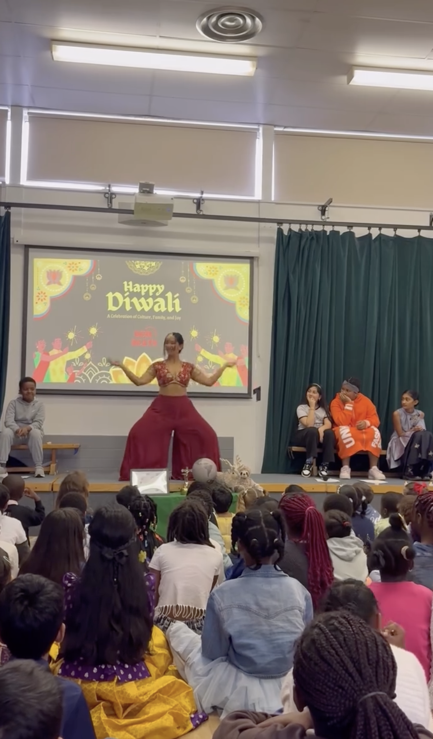Children and a performer on stage celebrating Diwali with a slideshow on the screen that says "Happy Diwali" and depicts traditional motifs and children celebrating.