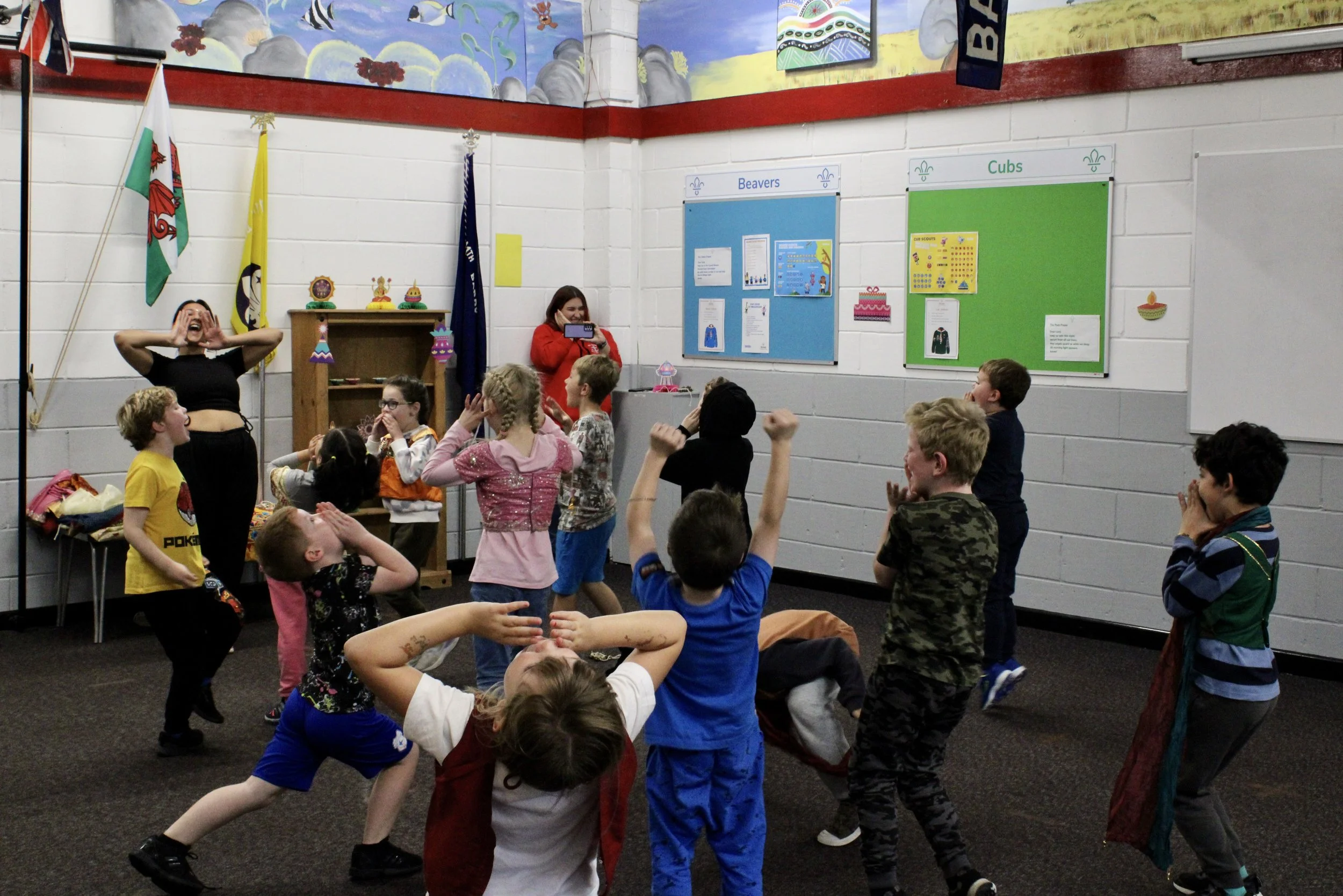 Children participating in a group activity in a classroom, with two women leading the activity. The classroom walls feature educational posters and colorful decorations, including flags and artwork.
