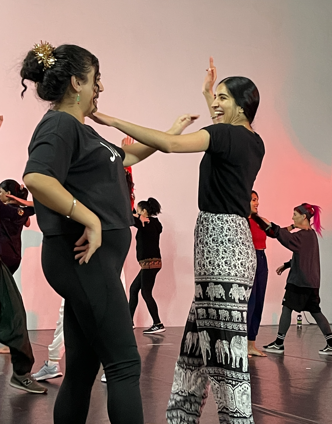 Two women smiling and dancing together, with multiple people dancing in the background on stage.