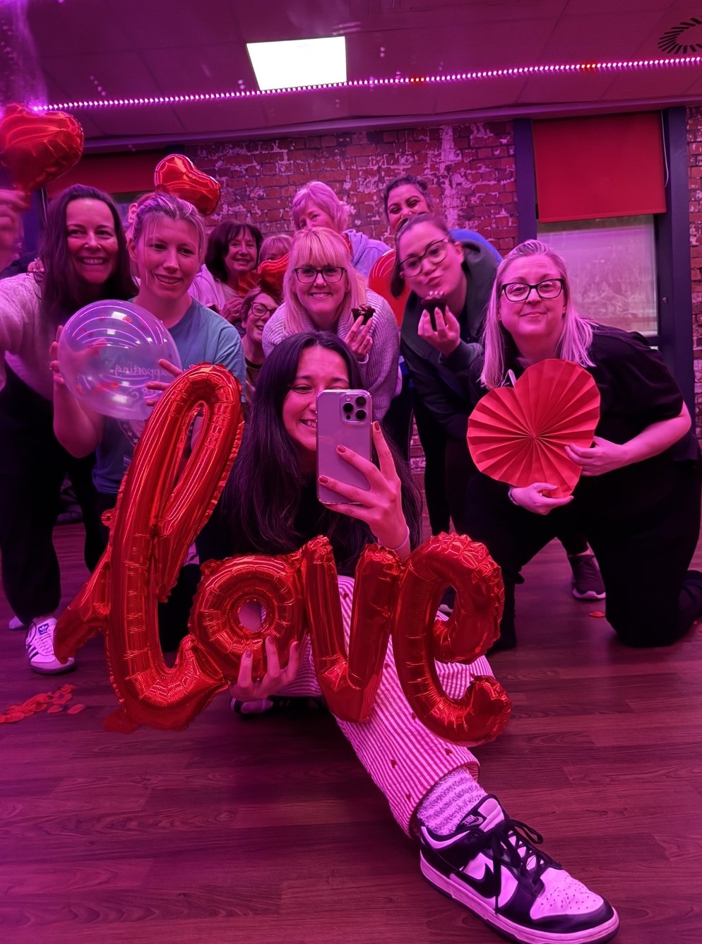 Group of women celebrating in a decorated room, some holding red heart-shaped balloons, a clear balloon, and red letter balloons spelling 'love', with a woman taking a selfie.
