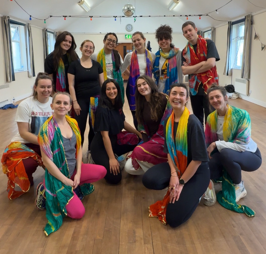 Group of eleven young women and one young man posing in a dance studio. Some women are sitting or kneeling, others are standing. All are wearing colorful scarves and smiles, with two women in tie-dye shirts and one man in a tie-dye shirt. The studio has windows, curtains, string lights, and a disco ball hanging from the ceiling.