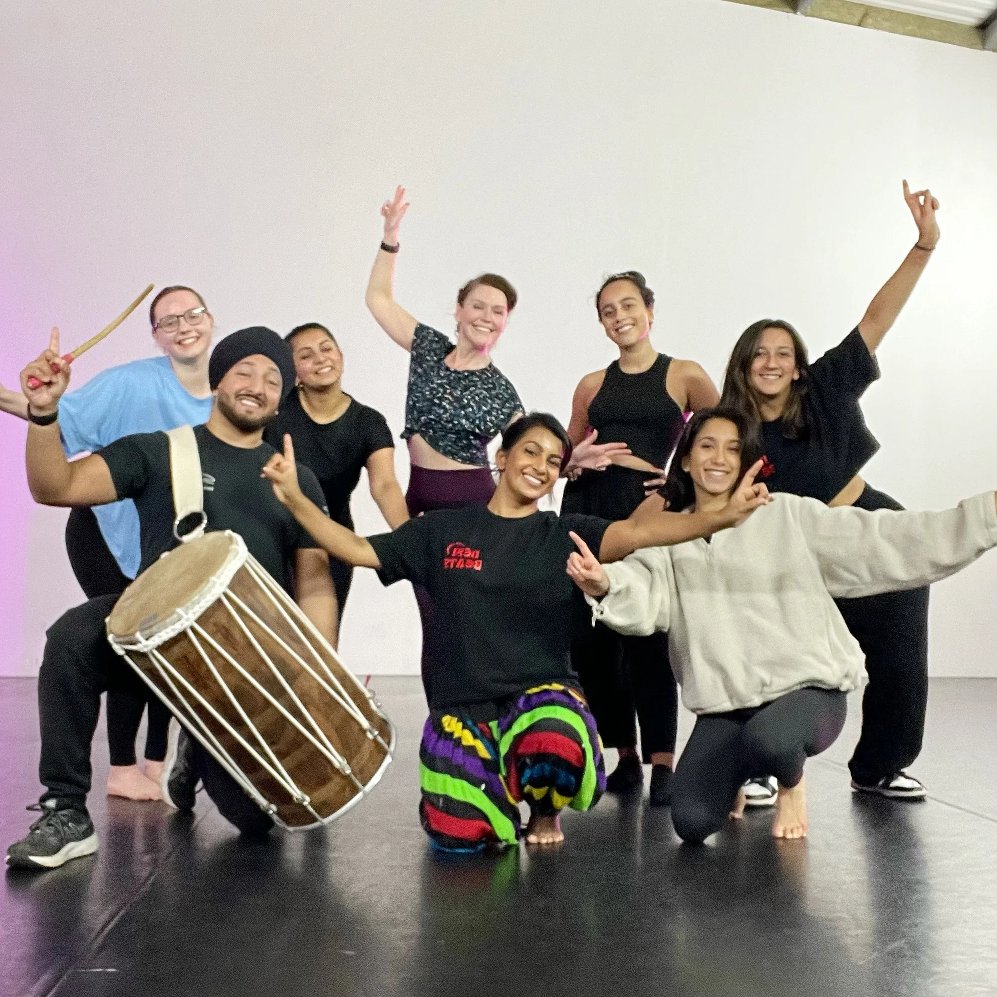Group of smiling people posing together, some holding a drum and drumsticks, in a dance or performance studio with plain white walls.