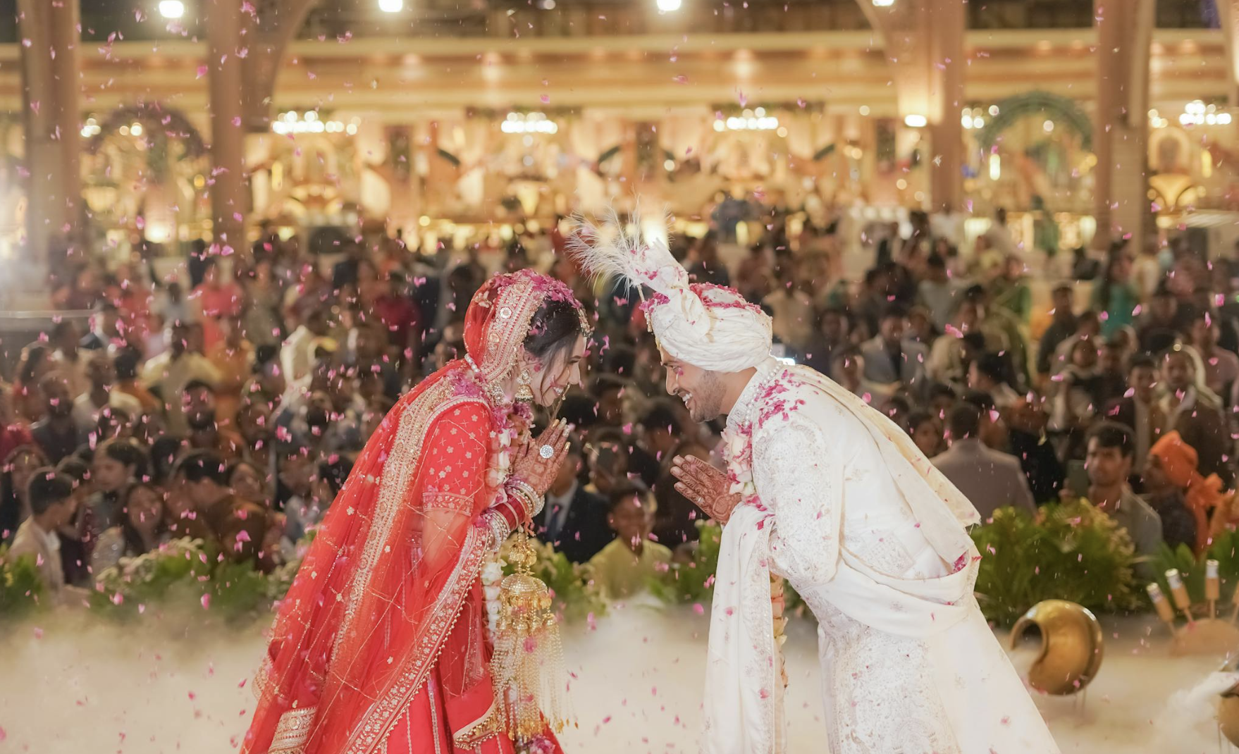 A bride and groom dressed in traditional Indian wedding attire tilting forward with hands pressed together in a gesture of greeting or prayer, amid pink flower petals falling, in the middle of a large celebration with many guests in the background.