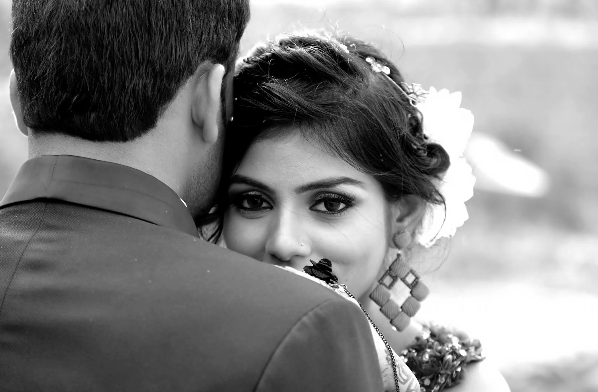 A black and white close-up photo of a woman with dark hair and earrings, looking directly at the camera, with her face partially hidden behind a man’s shoulder.