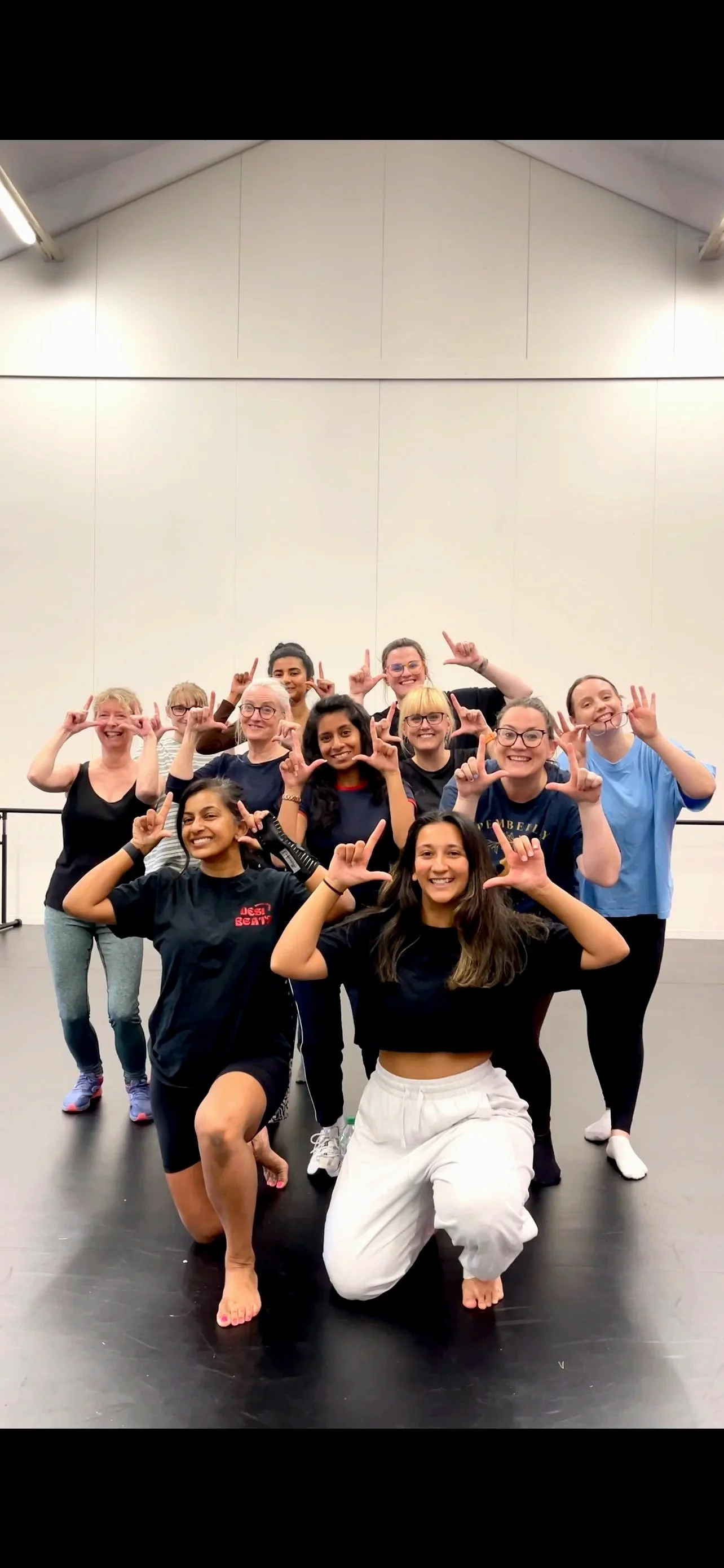 A group of women posing together in a dance studio, smiling and making hand gestures, some in casual clothing and some in dance attire.