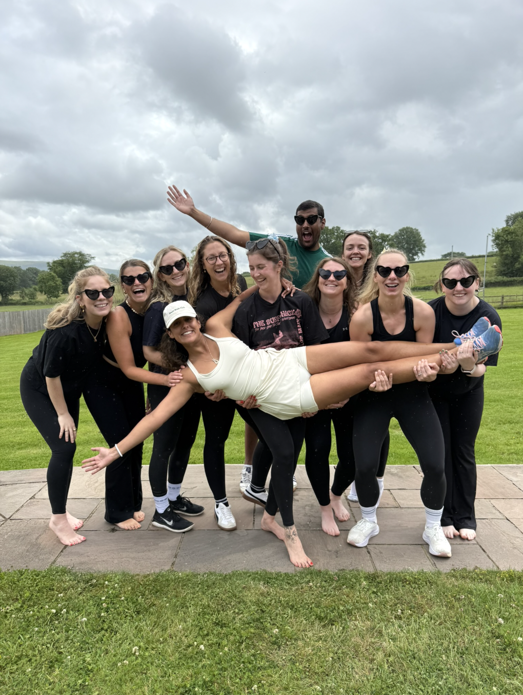 Group of women in athletic clothing smiling and holding a woman horizontally, outdoors on a cloudy day.