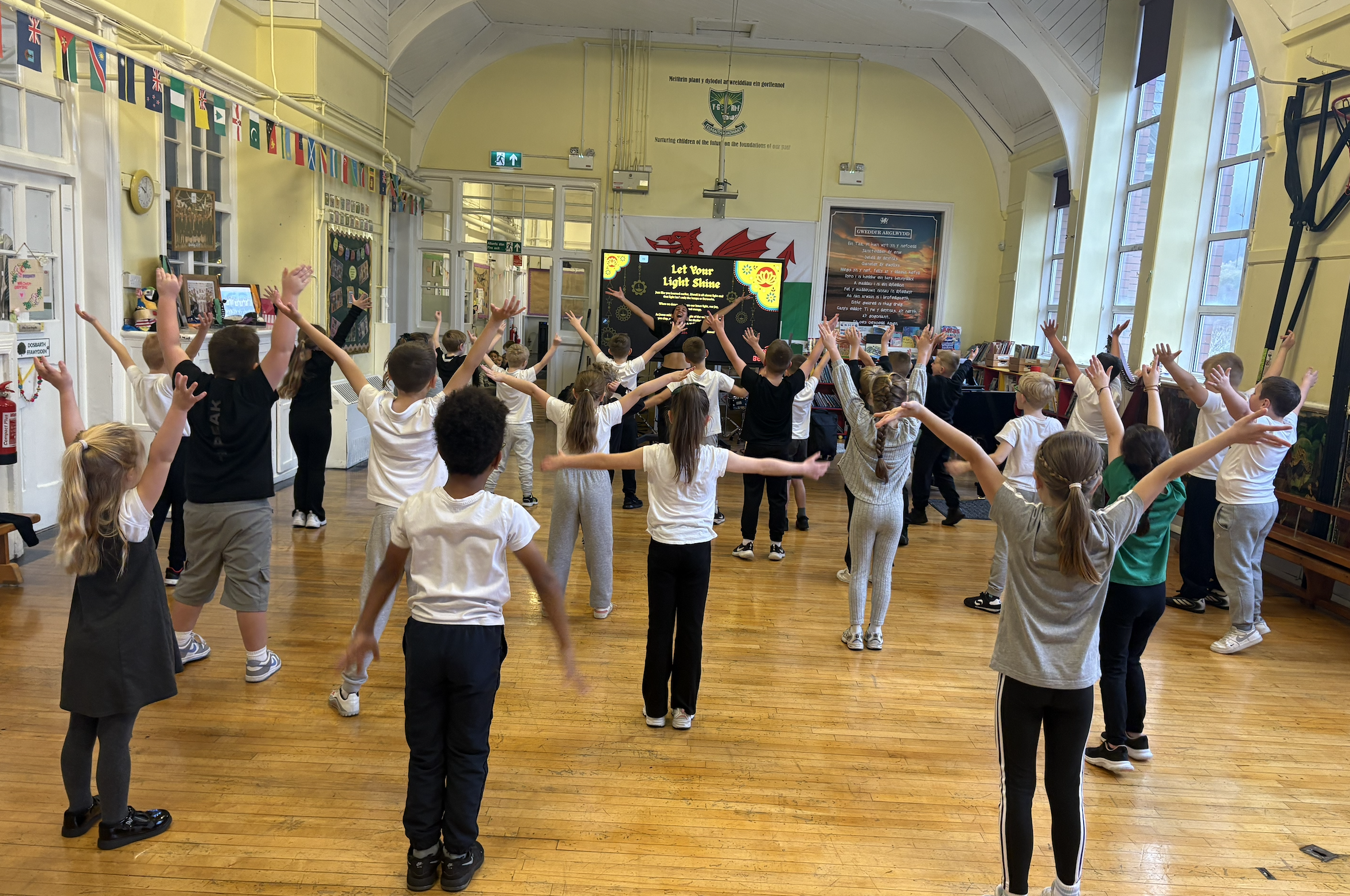 Classroom with children practicing a dance or exercise routine, facing a screen with a colorful display, and arms raised in the air.