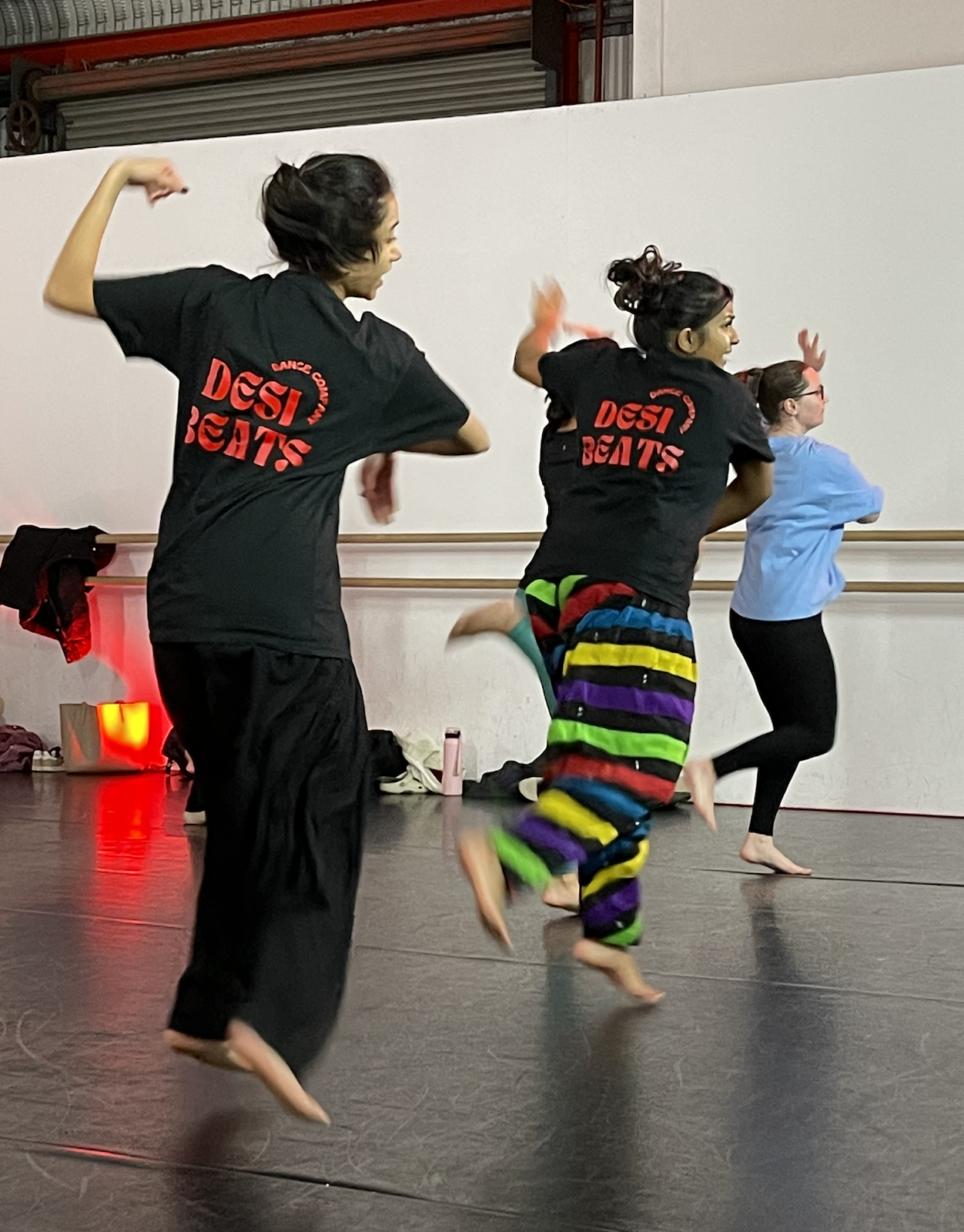 Three women practicing dance or fitness in a studio, two wearing black 'Desi Beatz' shirts, one in colorful striped pants and the third in black leggings and light blue shirt.