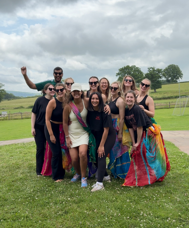 Group of people outdoors on a grassy field with cloudy sky, some wearing colorful skirts and others in black clothing and sunglasses, celebrating.