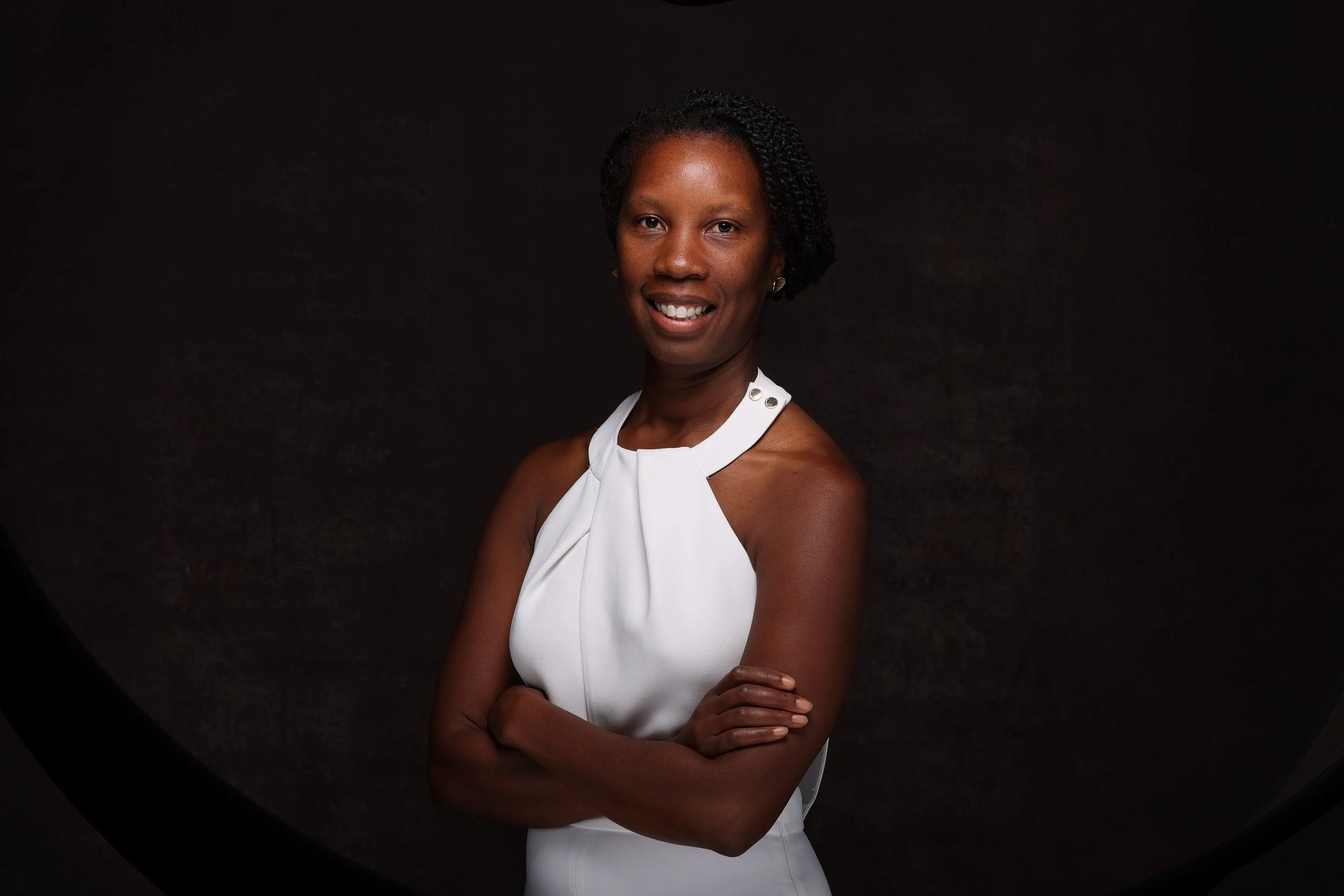 A woman with dark skin and short curly black hair, wearing a sleeveless white dress with a high neckline, standing against a dark background and smiling with arms crossed.
