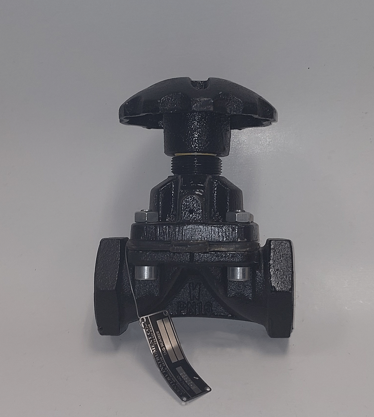 A black industrial valve with a large handle at the top, mounted on a white surface, with a tag attached to its base.