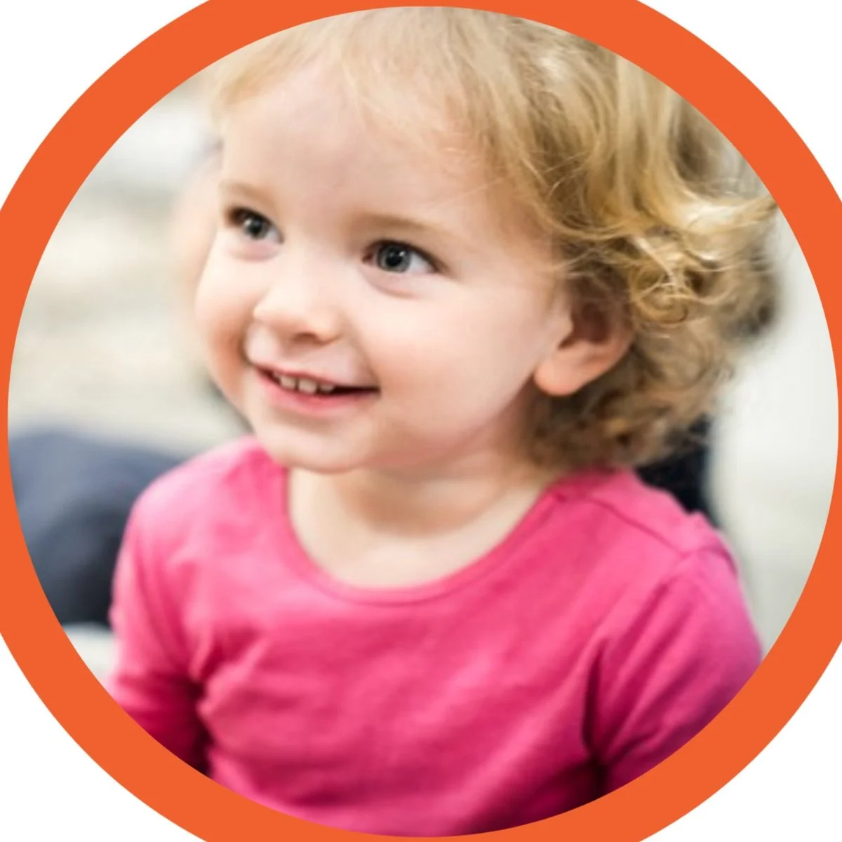 A young girl with curly blond hair and blue eyes wearing a pink shirt, smiling and looking to her left.
