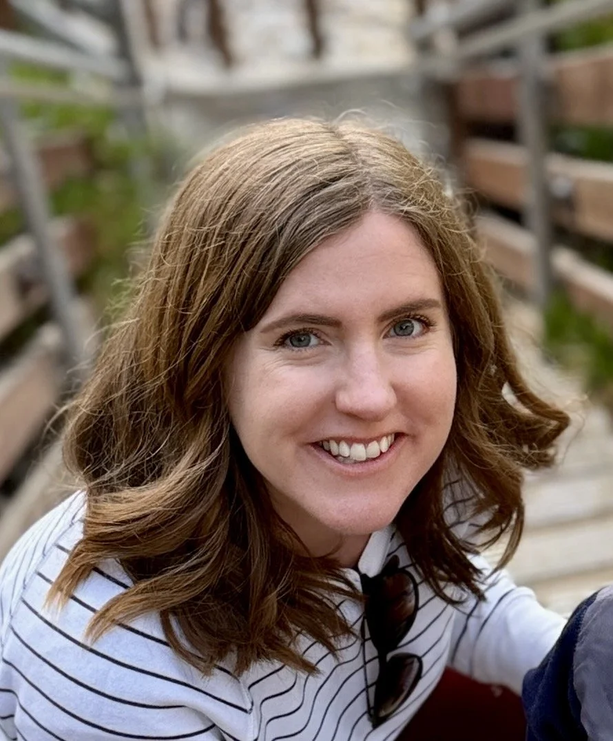 A woman with shoulder-length wavy brown hair smiling outdoors, wearing a white and black striped shirt.