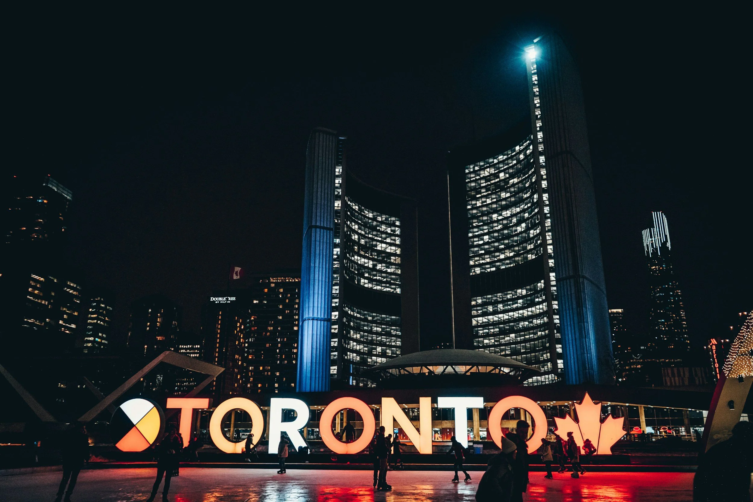 Night scene of downtown Toronto with illuminated Toronto sign featuring a red maple leaf, people walking, and tall city skyscrapers, including the CN Tower in the background.