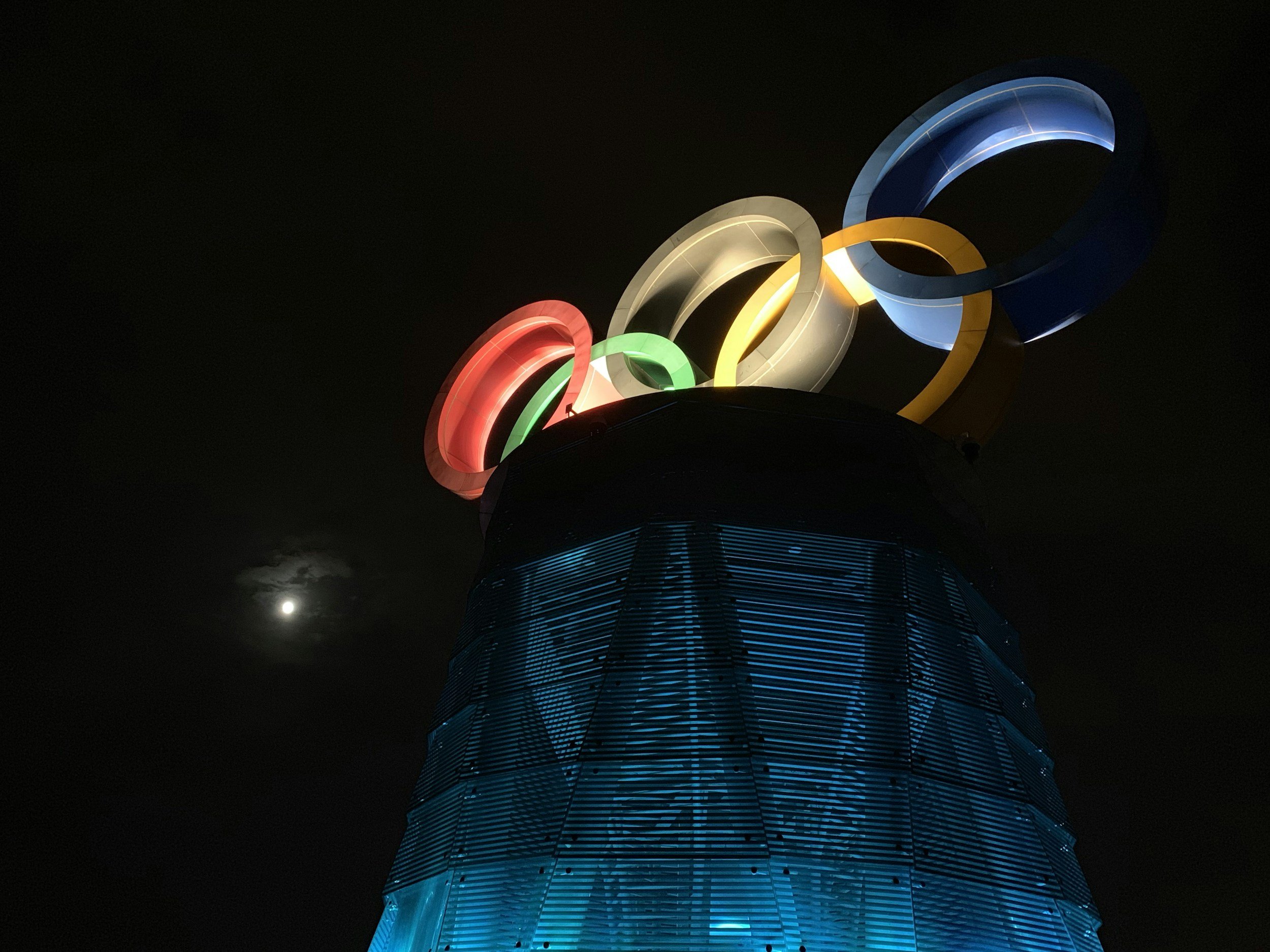 Nighttime view of the Olympic rings illuminated on a tall building, with the moon visible in the dark sky.