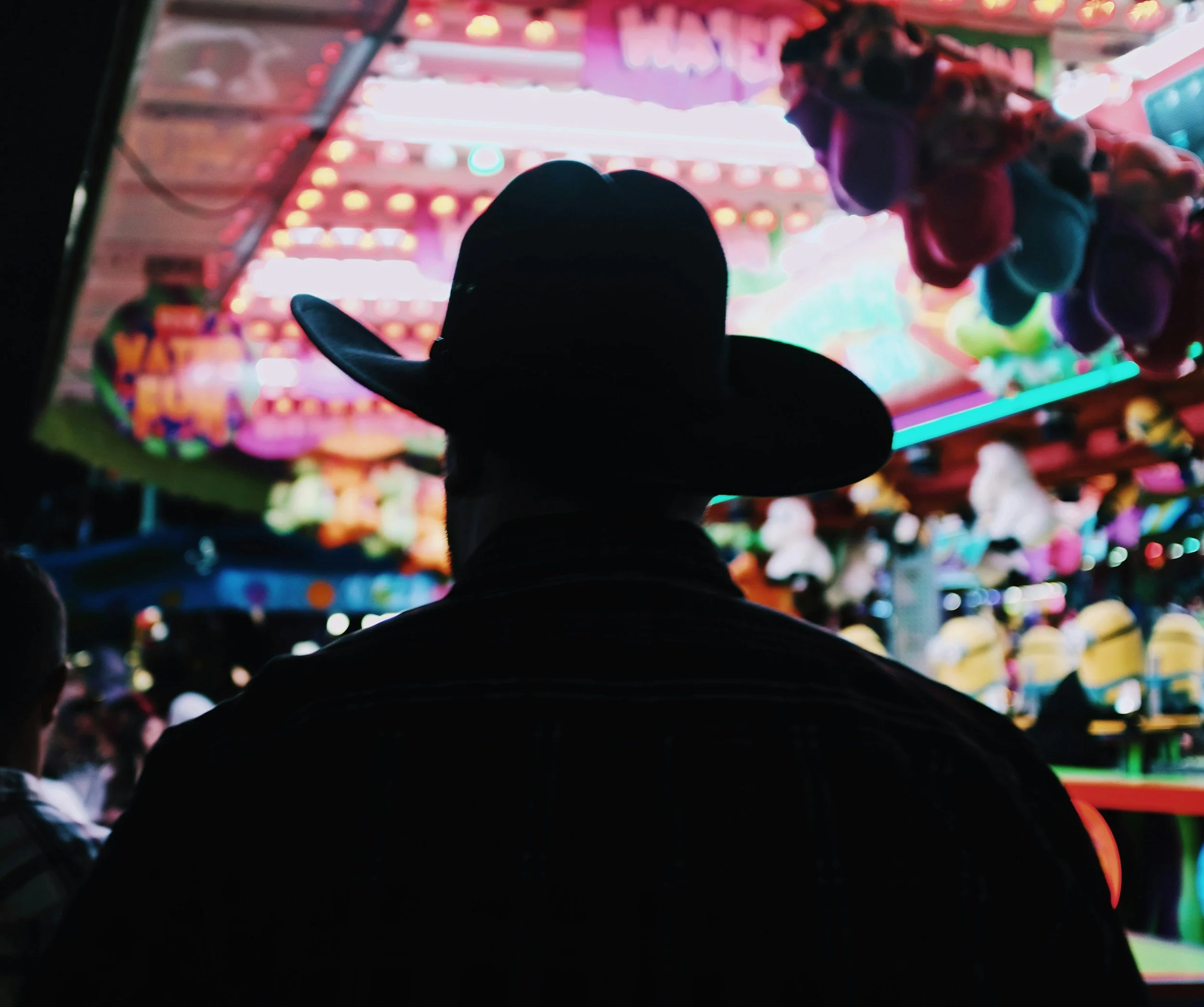 A person wearing a cowboy hat standing in front of colorful arcade game machines at a carnival or fair.