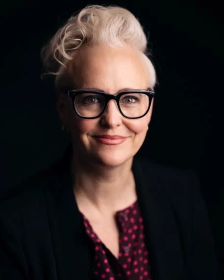 Portrait of a woman with short white curly hair, glasses, and a black blazer, smiling against a dark background.