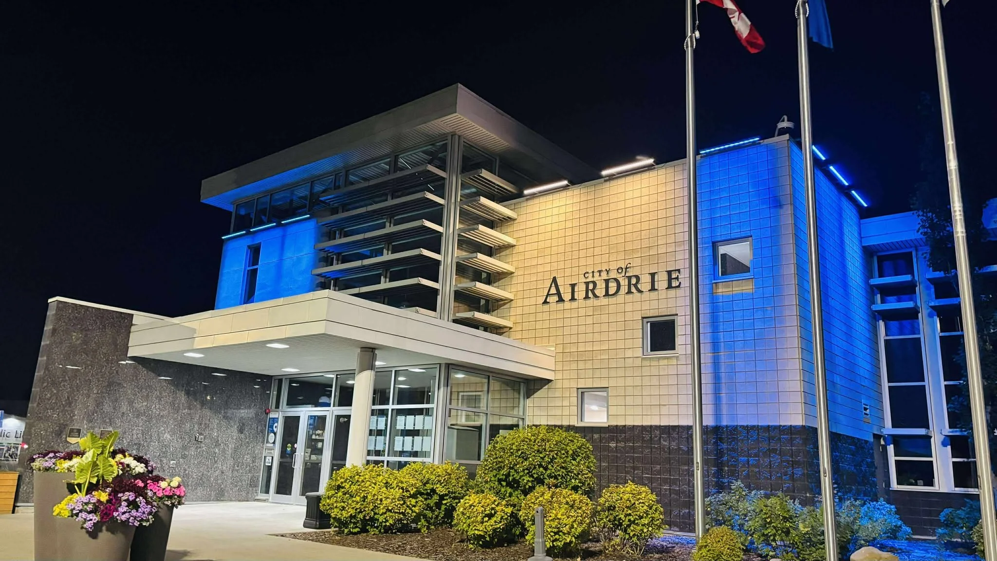 Night view of City of Airdrie government building with illuminated blue lighting, flags, and neatly landscaped bushes in front.