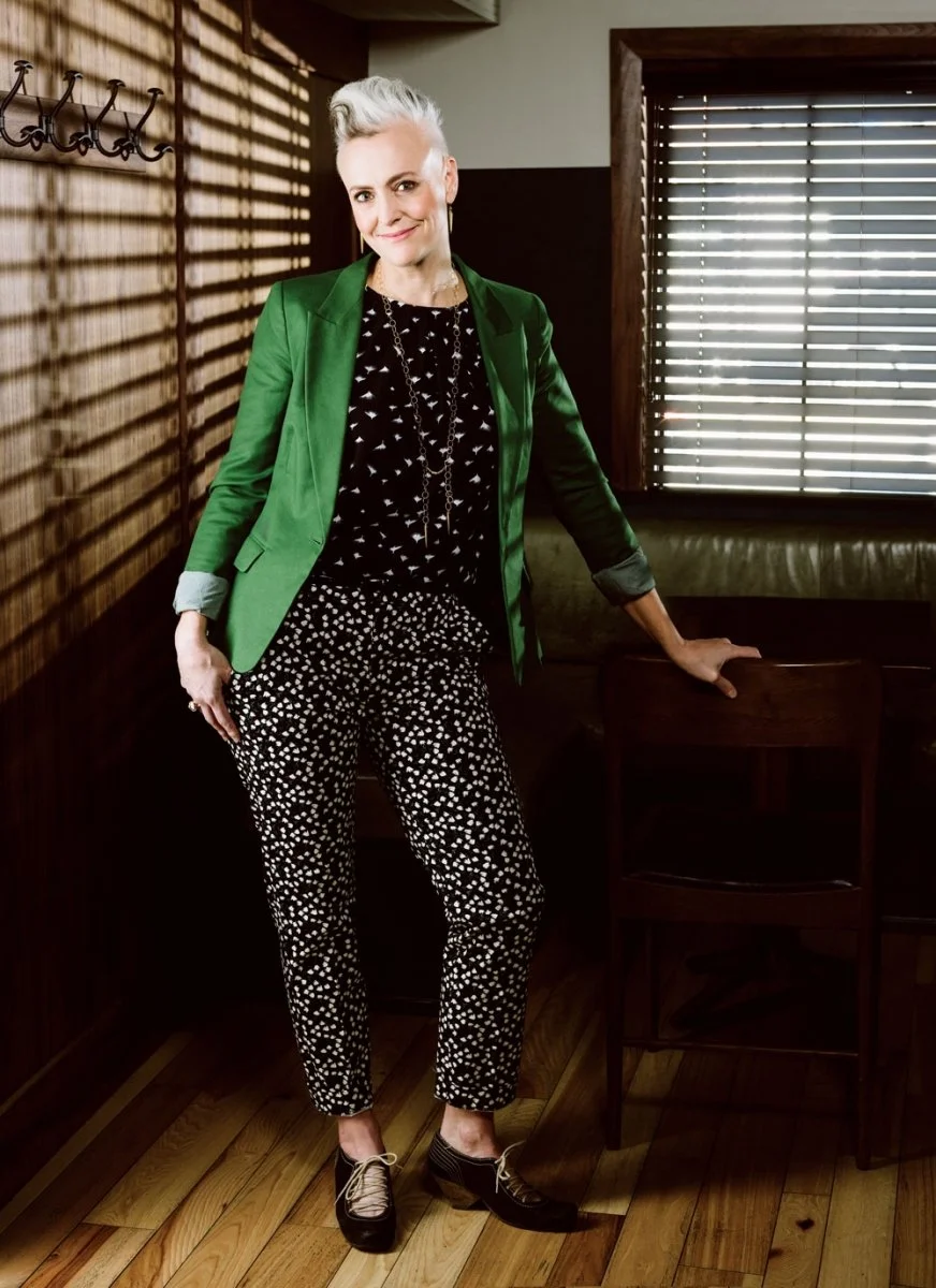 A woman with short gray hair and a confident smile, wearing a green blazer, black patterned top, black and white dotted pants, and black shoes, standing in a room with wooden walls and blinds on the window.