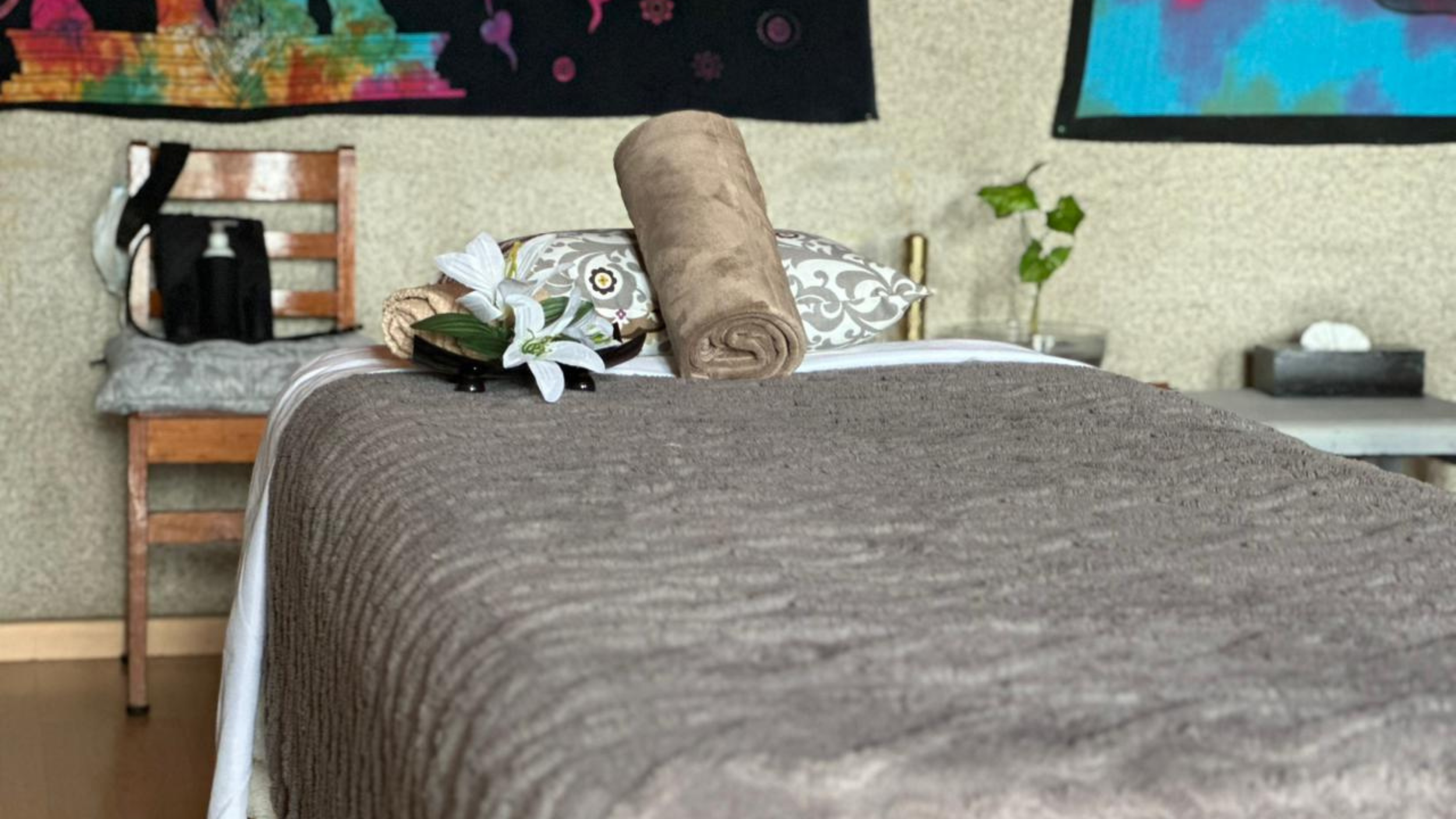 Cama arreglada con almohadas decorativas, un rollo de toalla, flores y un plato con flores blancas, en una habitación con paredes de textura y obras de arte.