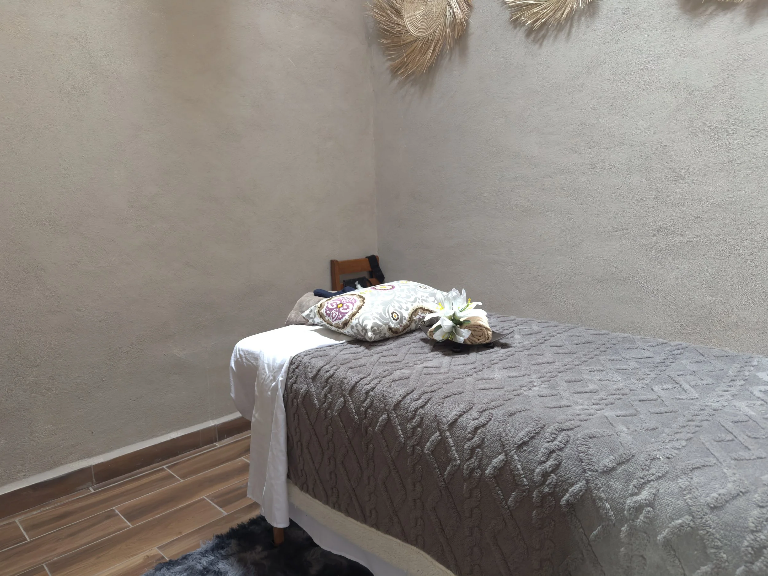 Cama sencilla en una habitación con paredes de textura ligera y piso de madera. La cama tiene un cubrec quien, cojines y flores blancas decorativas.