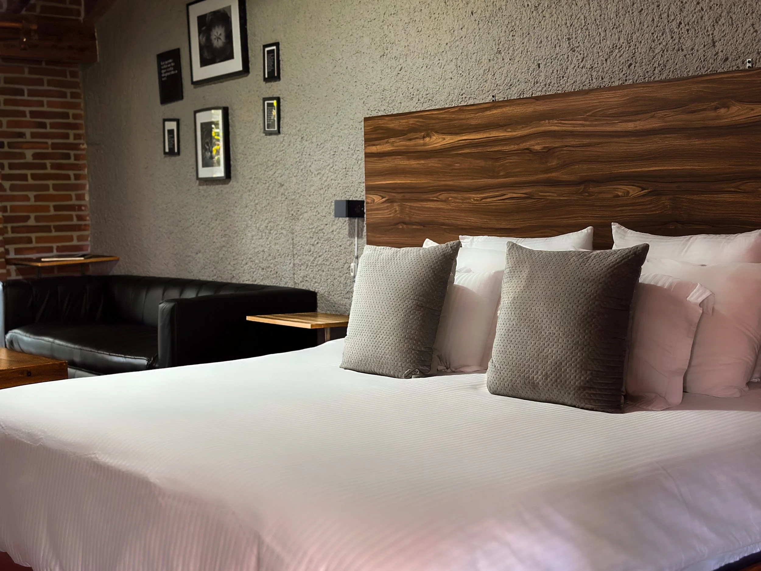 Cama con sábanas blancas y almohadas grises en una habitación moderna con pared de piedra y cuadros decorativos, un sillón negro de cuero y mesas de madera.