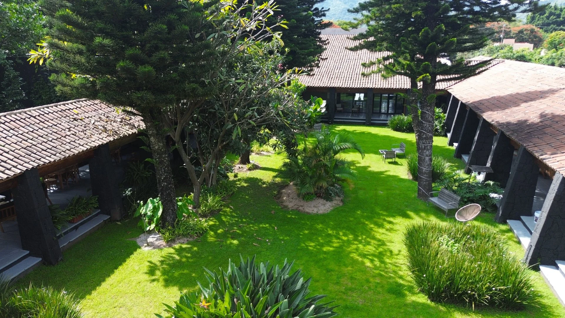 Jardín con césped verde, árboles grandes y pequeños, y casas con techos de teja roja en un entorno natural y soleado.