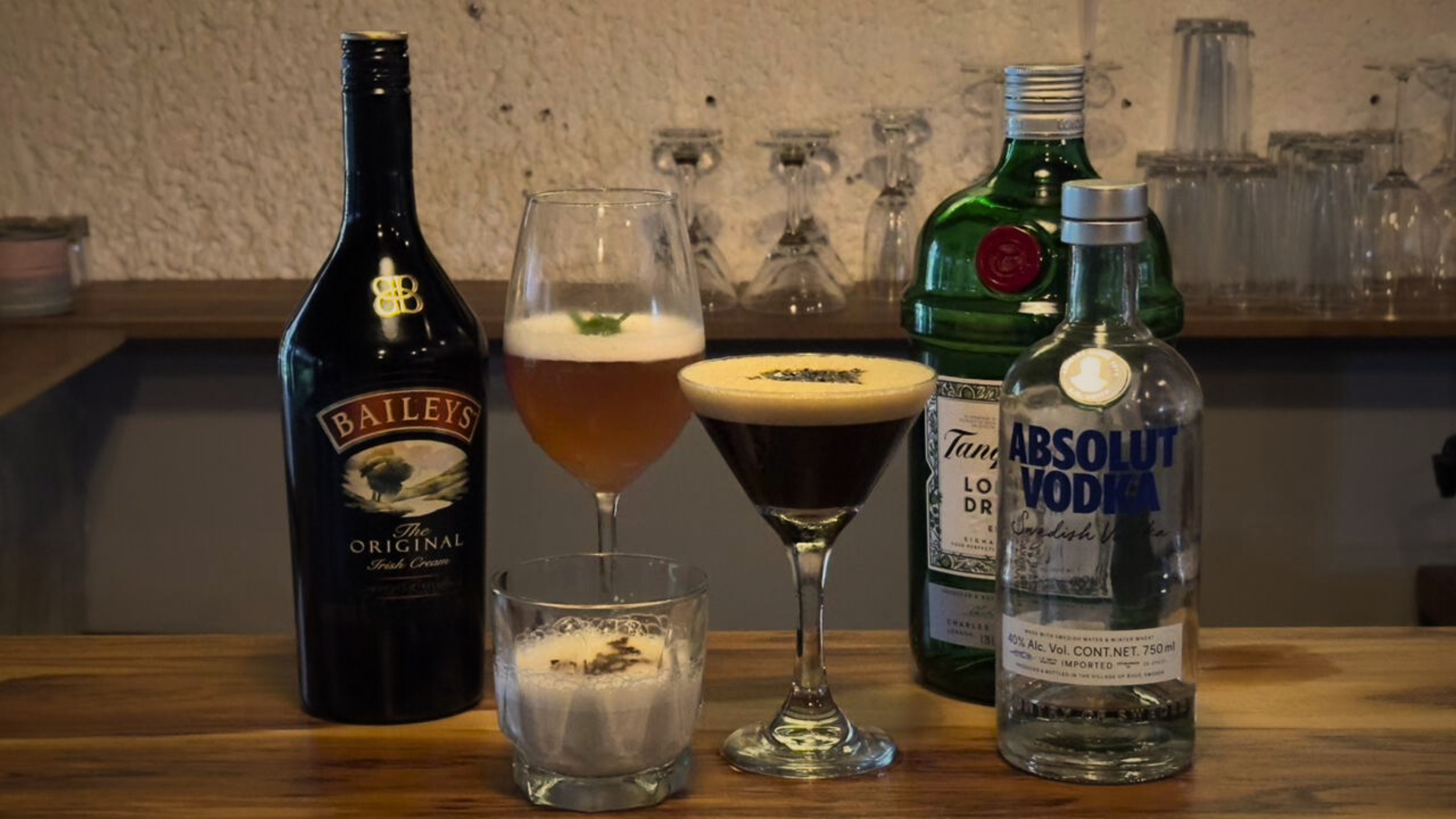 Variedad de bebidas alcohólicas y cócteles en una barra de madera, incluyendo una botella de Baileys, una copa con bebida espumosa, dos cócteles con hielo, una botella de Tanqueray y una botella de Absolut VODKA, con copas y vasos en el fondo.