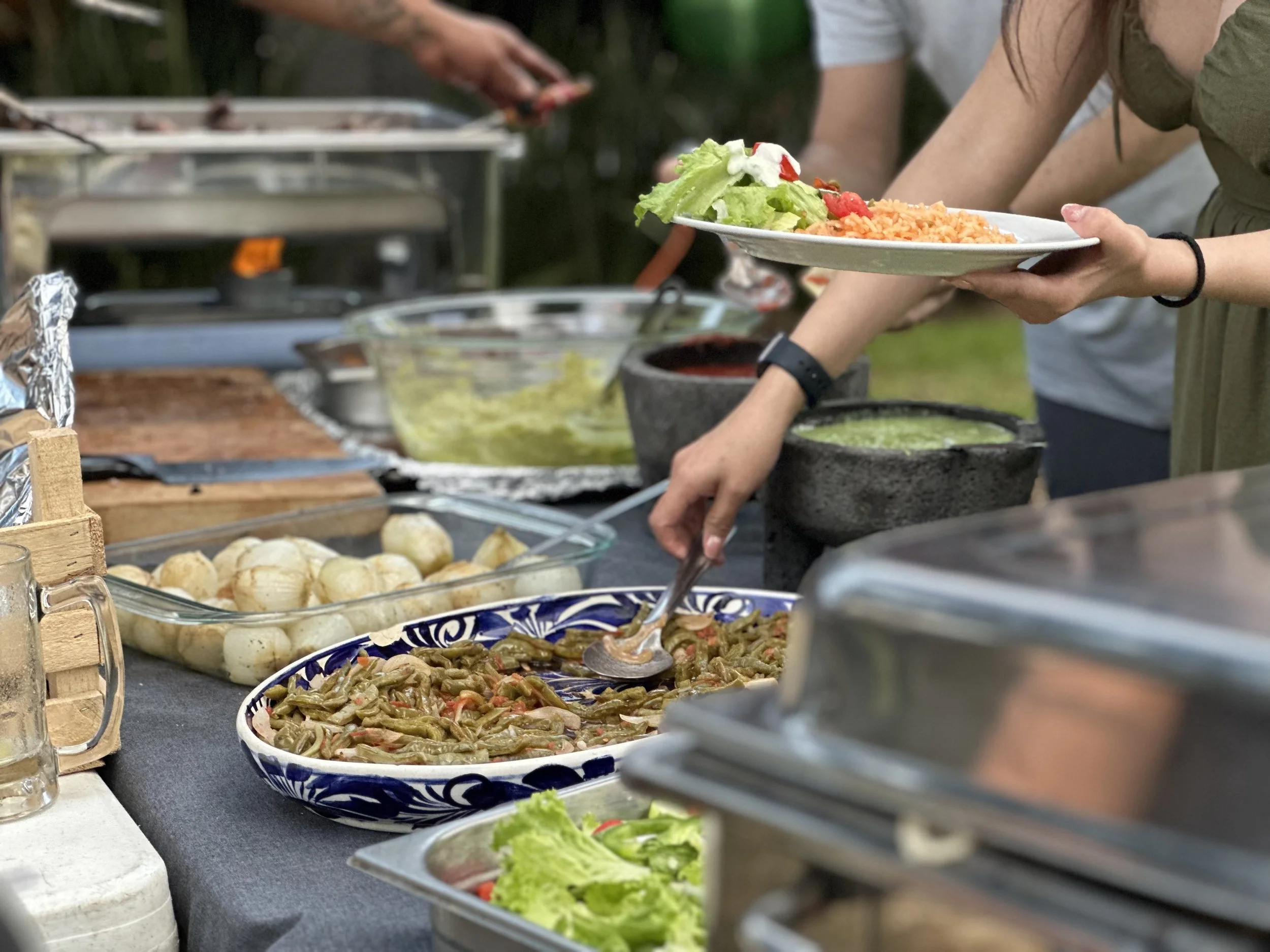 Personas sirviendo comida en una mesa en un evento al aire libre, con varios platos de comida como ensaladas, guisados y otros alimentos