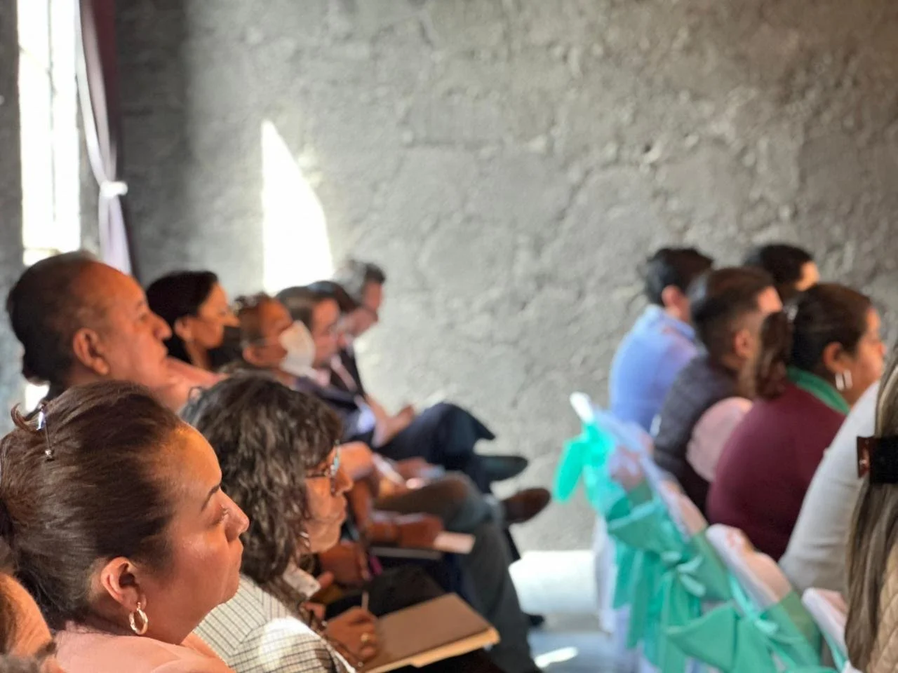 Diversas personas sentadas en fila en un evento, algunas con cuadernos o papeles, en un ambiente con pared de piedra y luz natural
