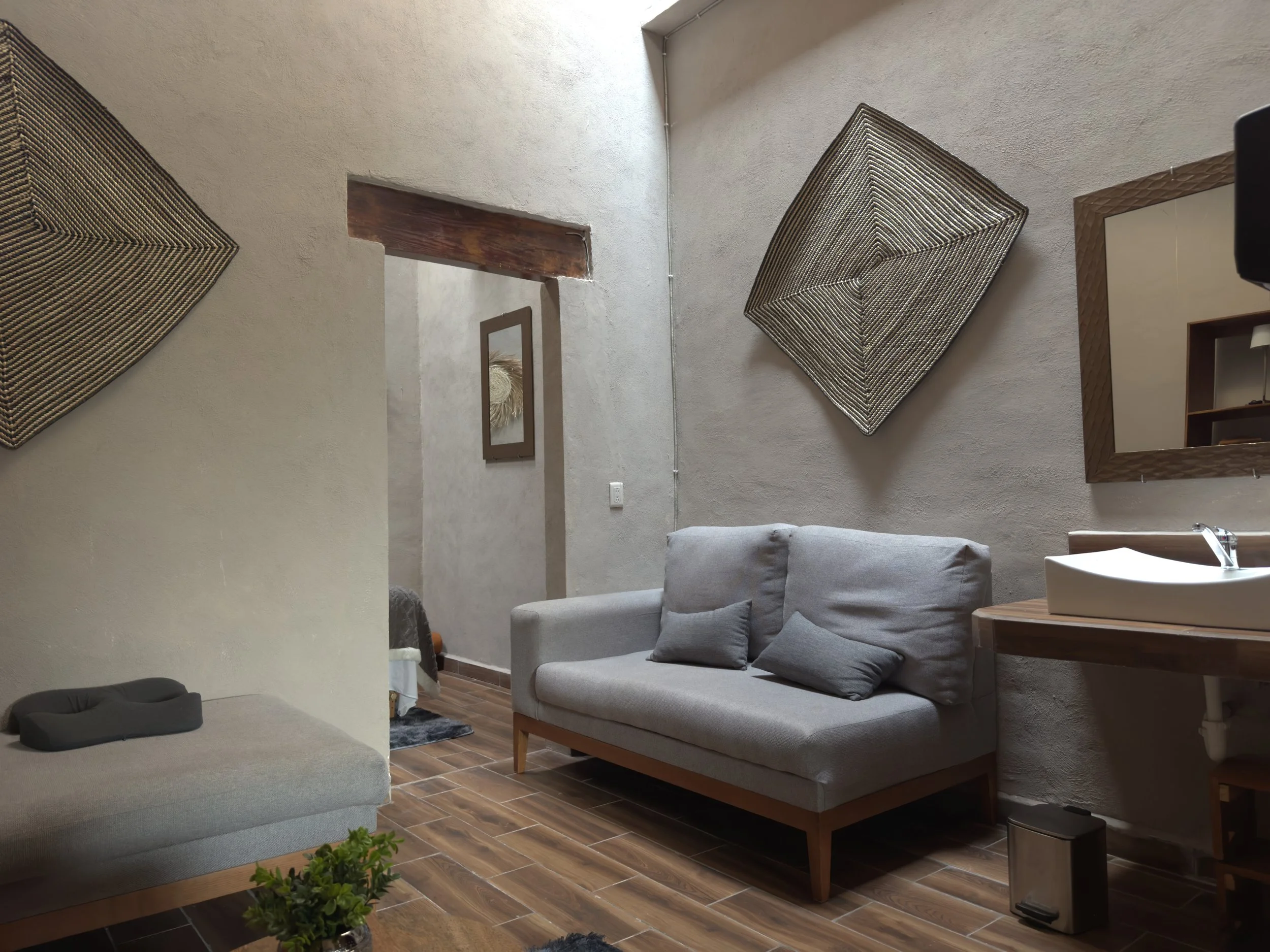 Sala de estar con sofá gris, cojines, espejo, decoraciones en la pared, lavabo y piso de madera.