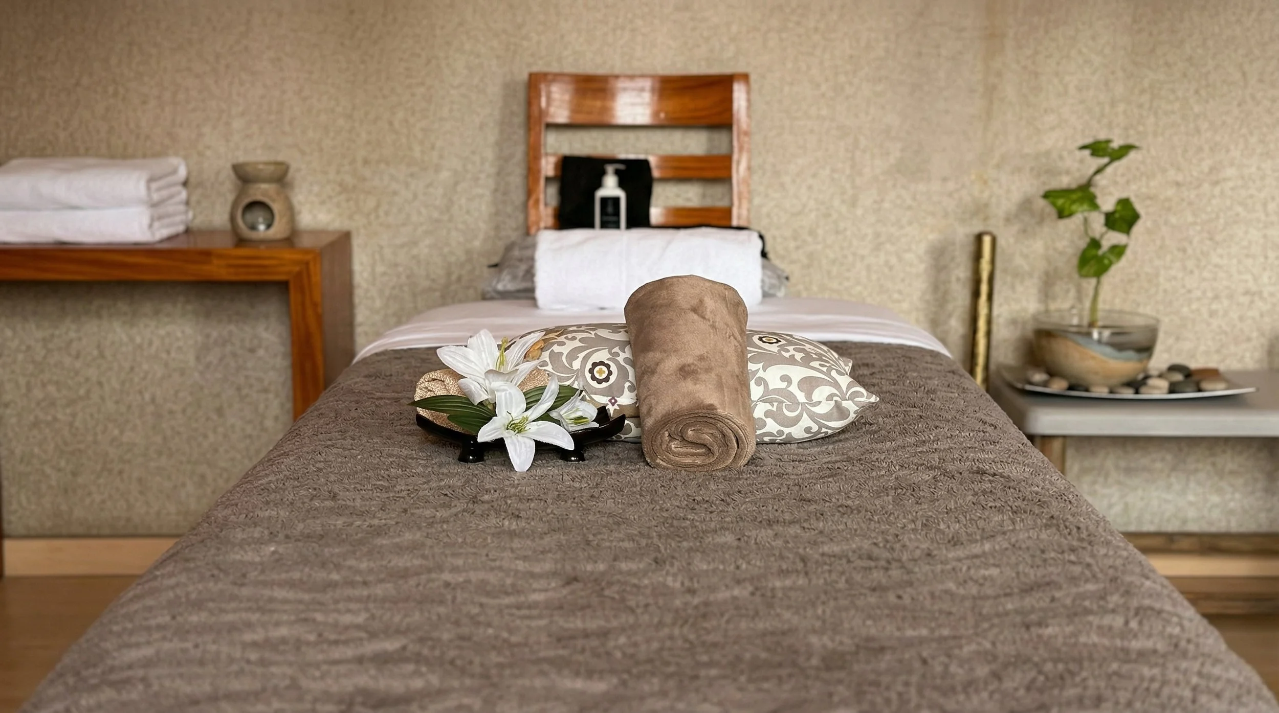 Cama arreglada con almohadas decorativas, un rollo de toalla, flores y un plato con flores blancas, en una habitación con paredes de textura y obras de arte.