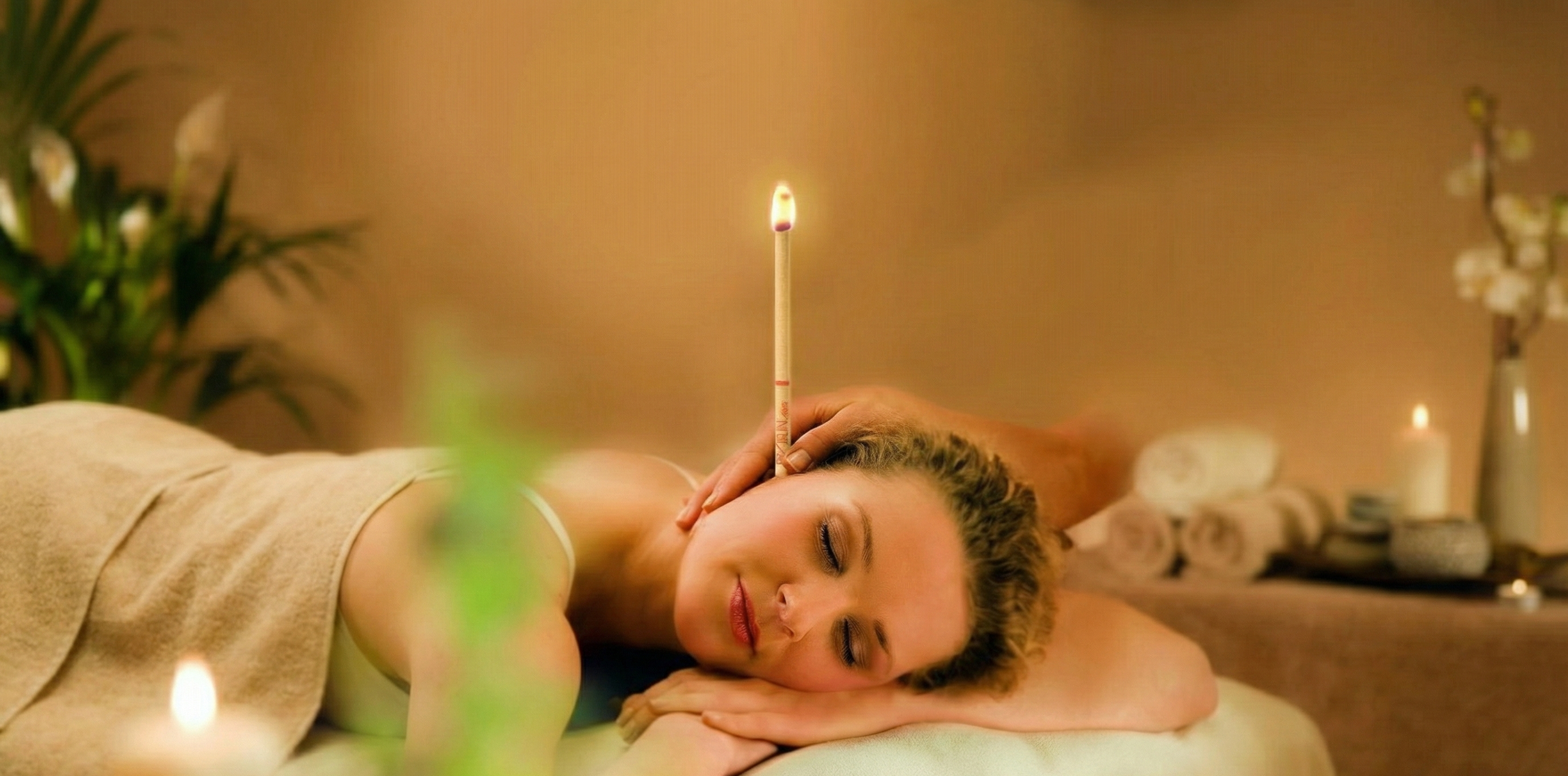 Mujer recibiendo un masaje, con una cerilla encendida equilibrada en la cabeza en un ambiente relajante con velas y plantas.