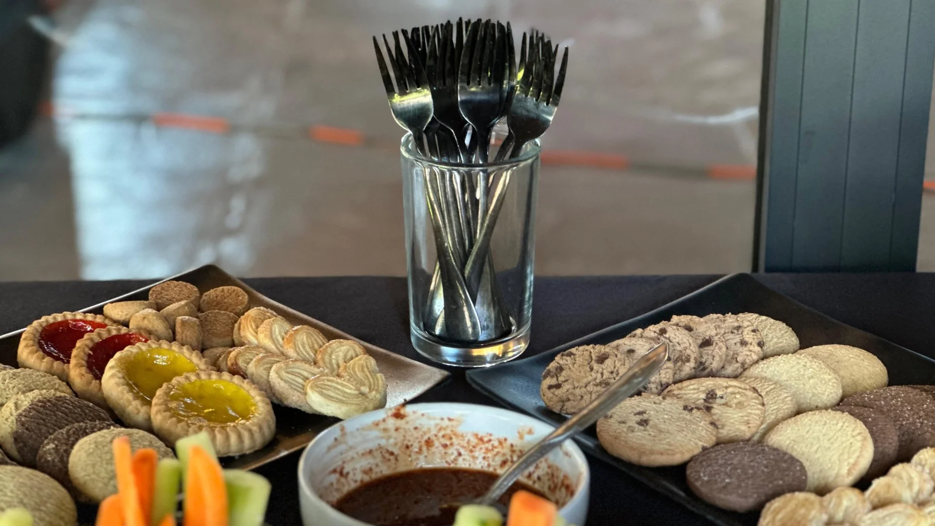 Variedad de galletas en platos negros y dulces en platos plateados, con un vaso con tenedores en el centro y un bol de salsa o miel en primer plano