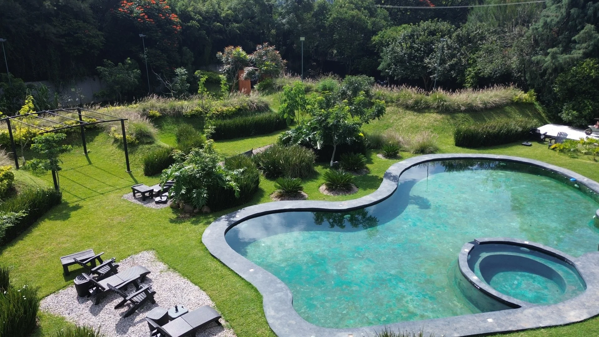 Piscina con forma orgánica en un jardín lleno de plantas, áreas de descanso y áreas de juego.