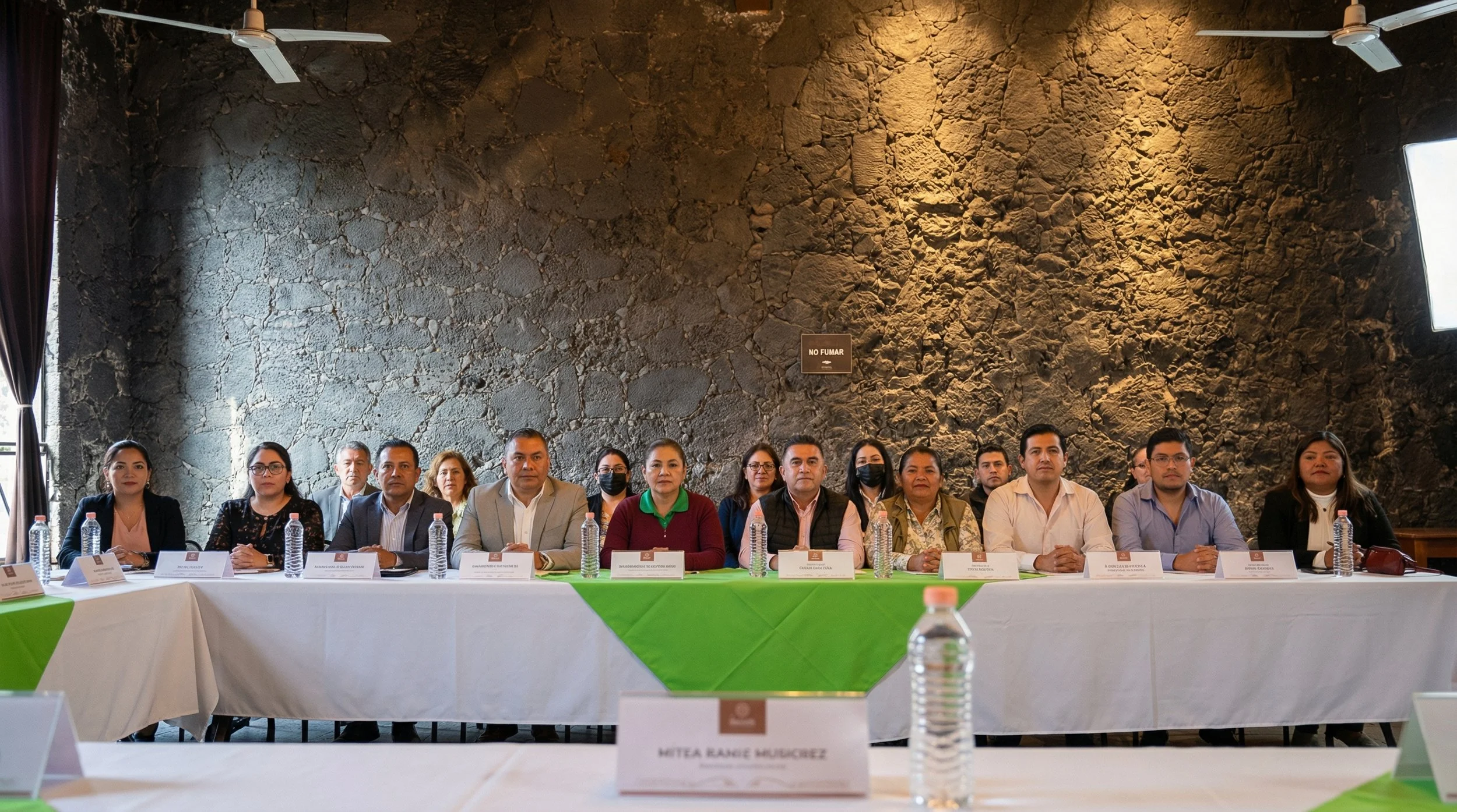 Reunión de varias personas sentadas en una mesa larga en una habitación con paredes de piedra oscuras y cortinas negras, algunas botellas de agua en la mesa, en un ambiente formal y de discusión.