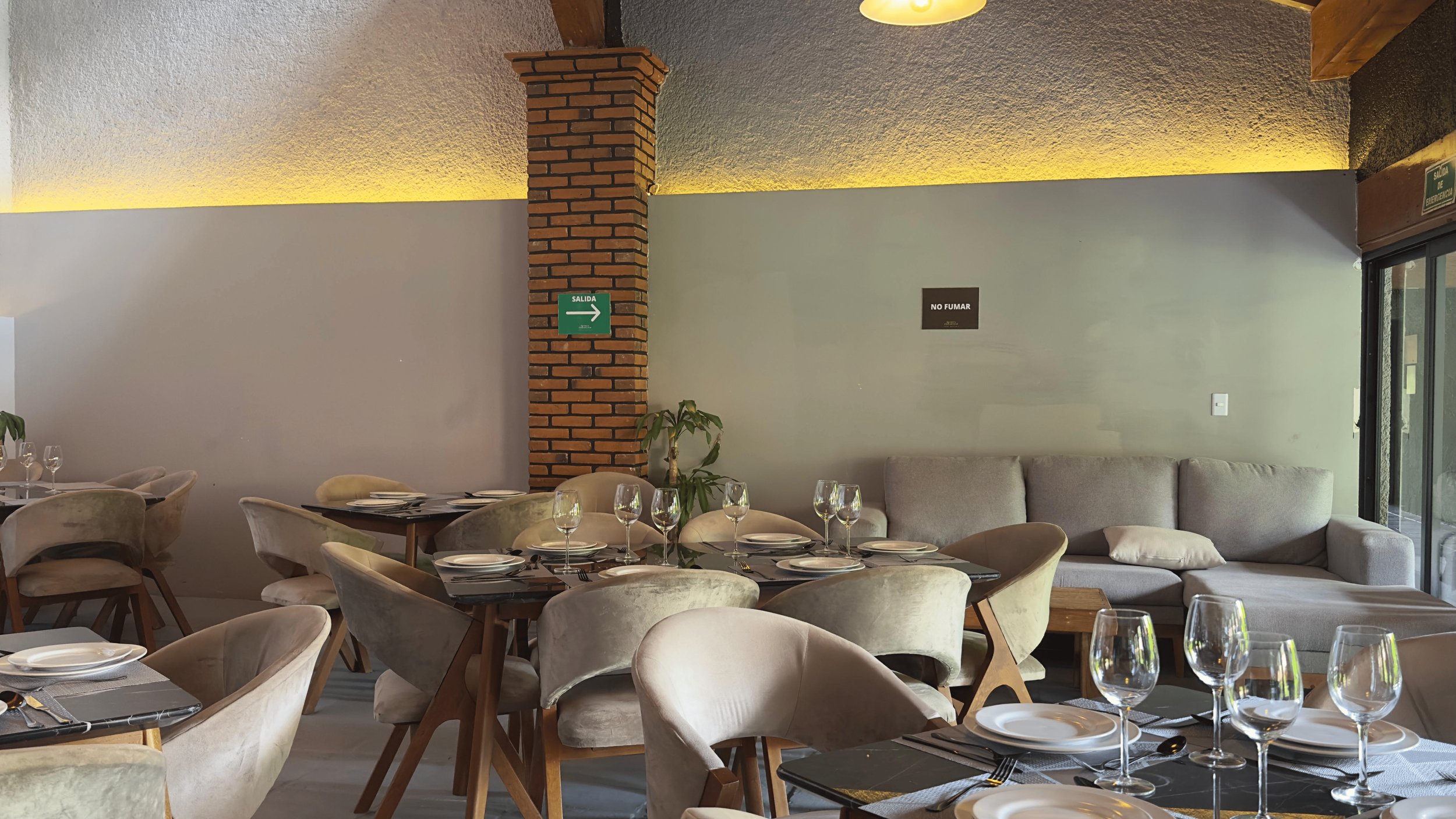 Sala de restaurante con mesas vestidas con platos y copas, sillones cómodos y una pared con señal de salida y cartel de no fumar.