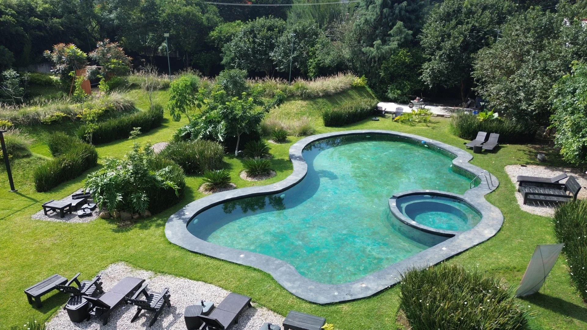 Vista aérea de una alberca en forma de corazón, rodeada de césped y suelo de piedra, con áreas de descanso y árboles en un entorno natural y verde.