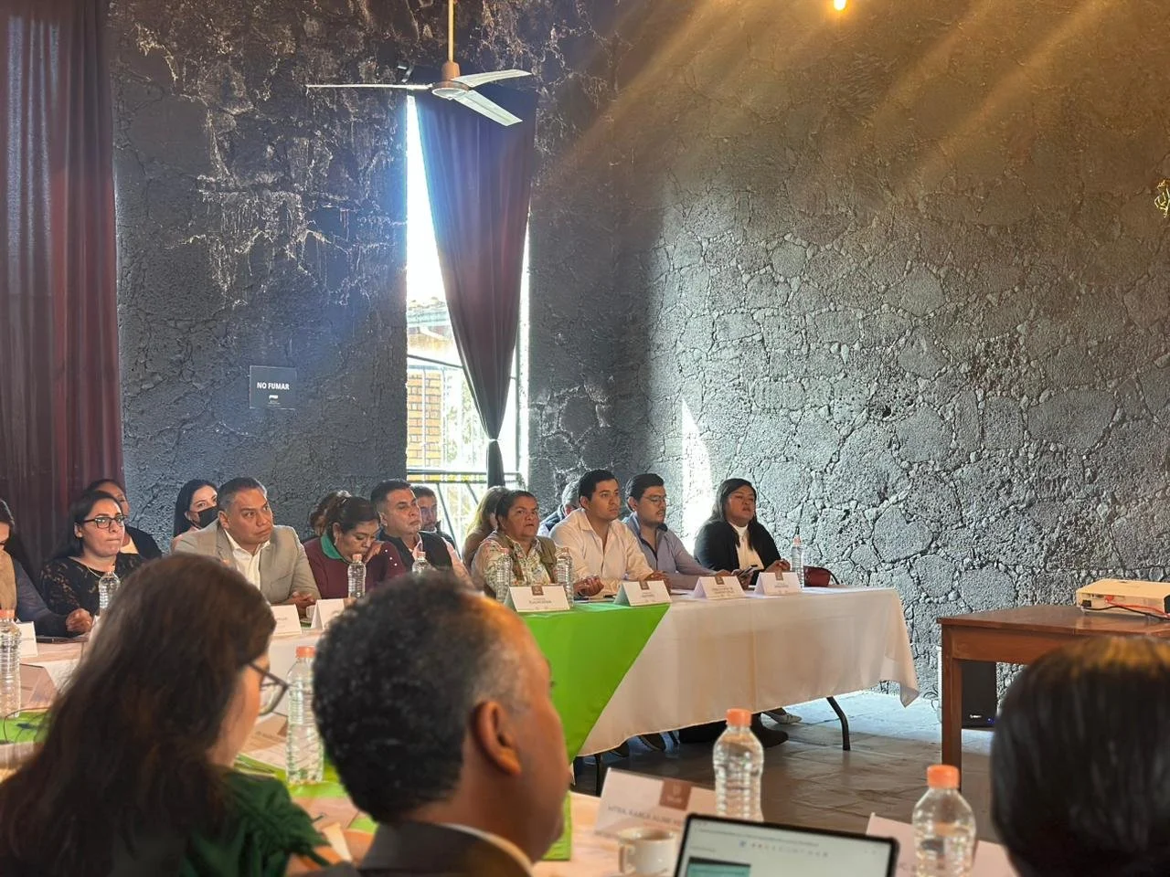 Reunión de personas sentadas en una mesa rectangular, en un ambiente interior con paredes de piedra y ventanas con cortinas, en una conferencia o reunión formal.