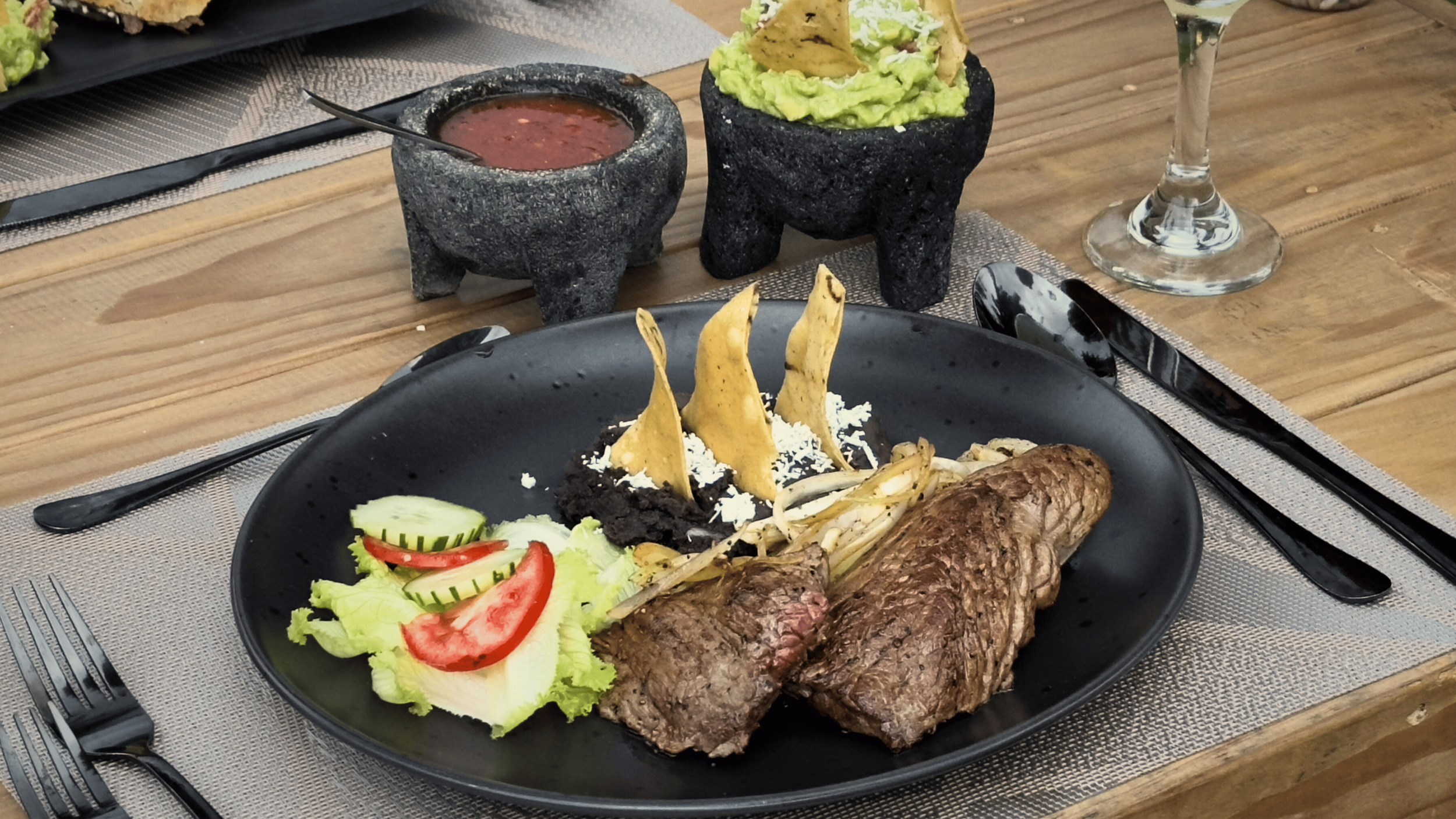 Plato con carne de res a la parrilla, guacamole, ensalada con tomate y pepino, y tacos dorados en un plato negro. En el fondo, dos molcajetes con salsa y guacamole, y una copa de vino.