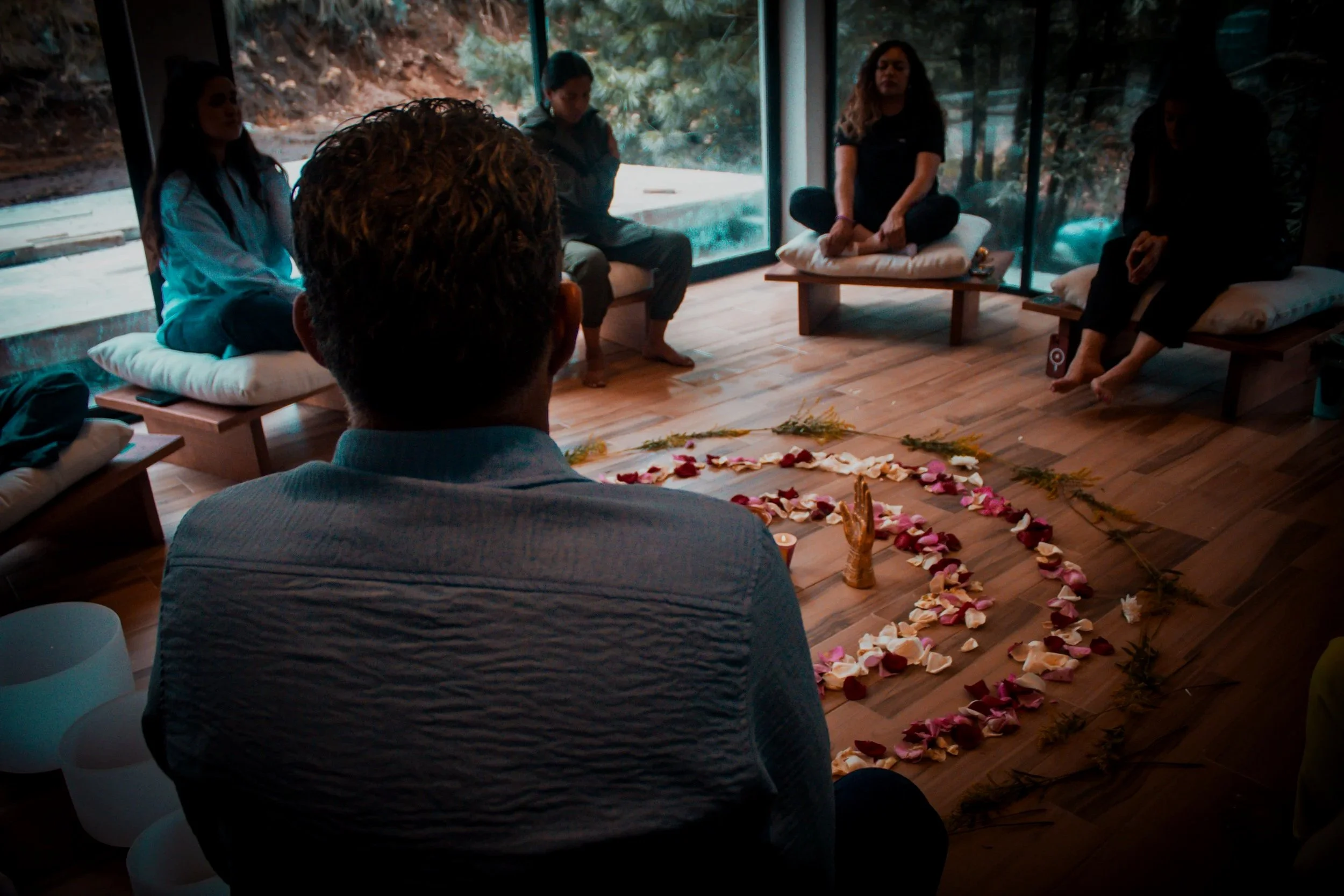 Grupo de personas participando en una ceremonia espiritual en un espacio interior con vista a un entorno natural, decorado con flores formando un corazón en el suelo.