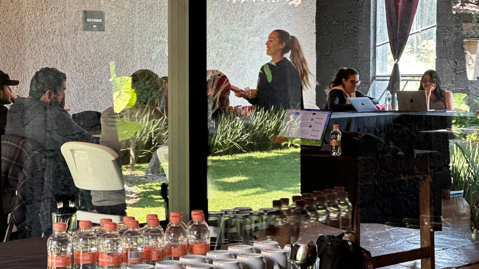 Personas en sala de reuniones vista a través de una ventana, con mesas y computadoras, y plantas en el jardín exterior.