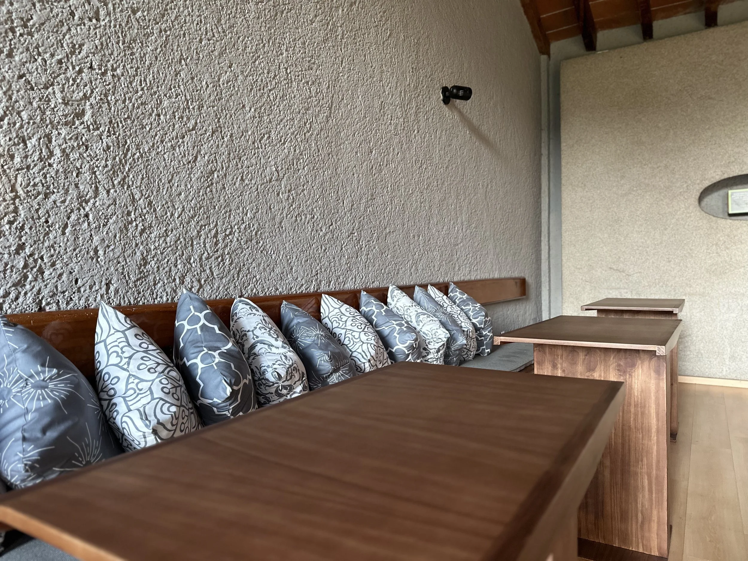 Sala de espera con varios cojines de diseño en un banco de madera contra la pared y mesas de madera al lado.