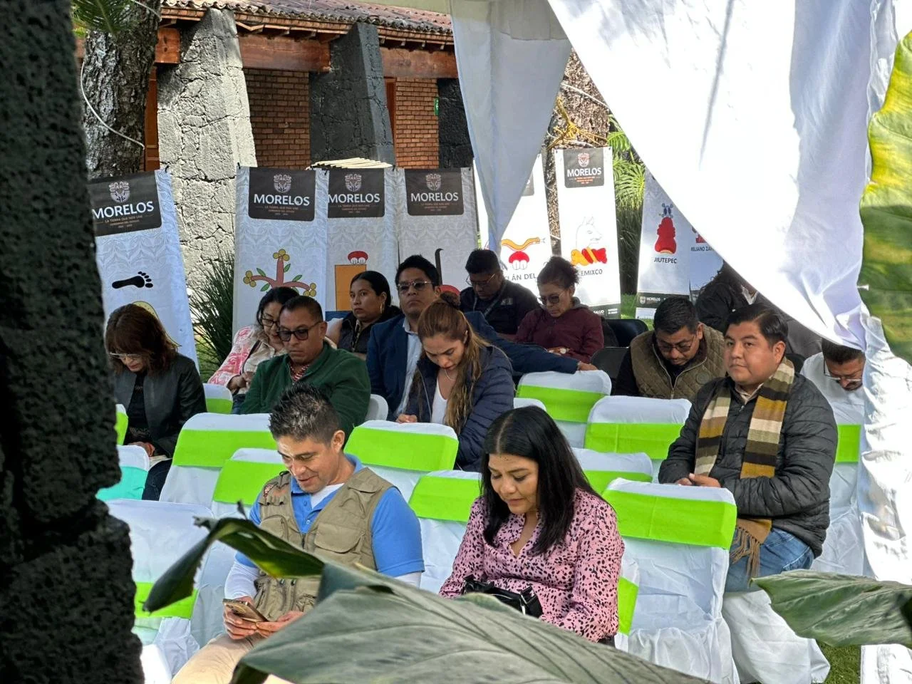 Reunión de personas en una ceremonia al aire libre entre árboles, con banners que dicen 'Morelos'.
