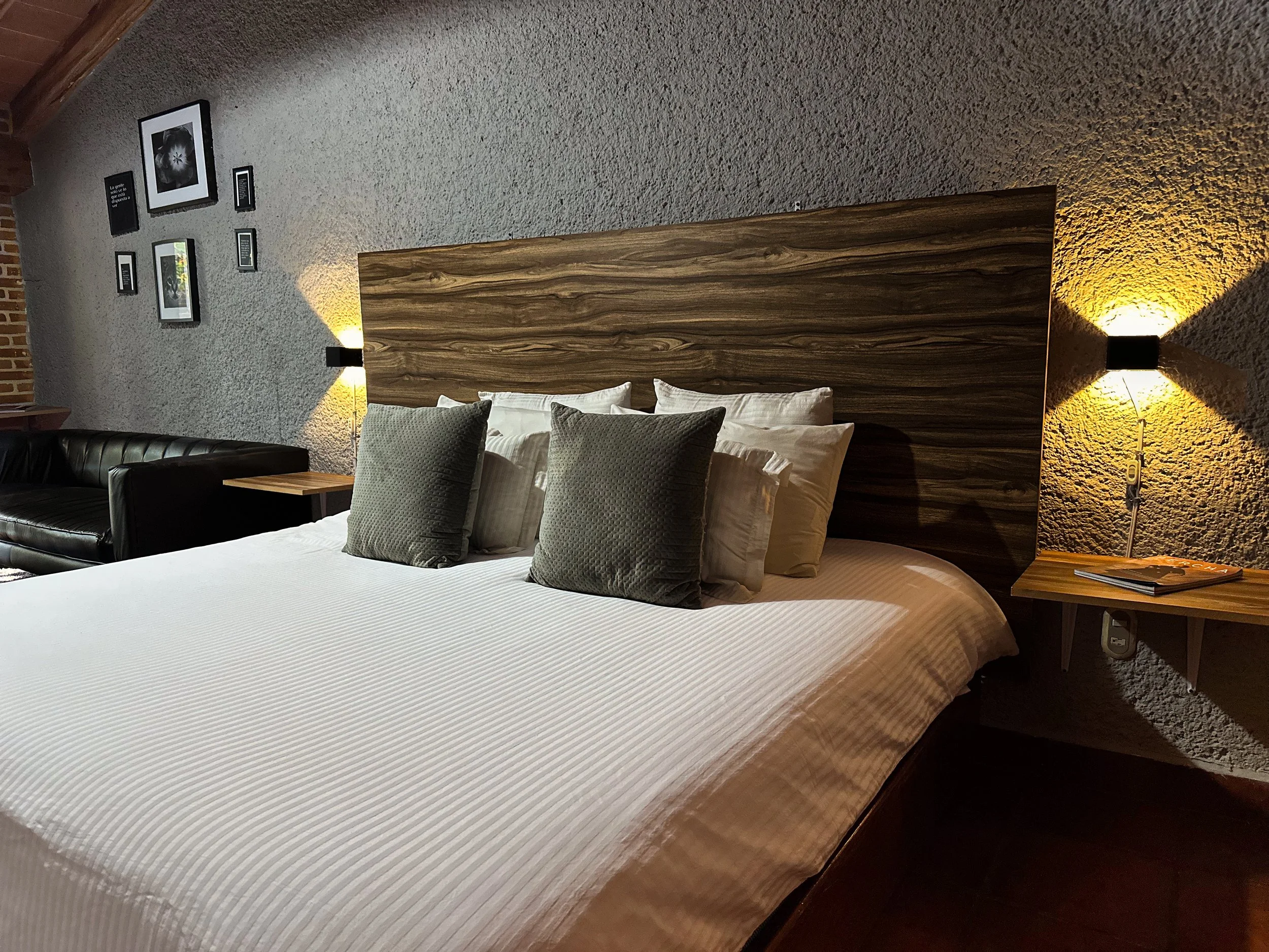 Cama doble con cabecera de madera, almohadas grises y blancas, lámparas de pared a cada lado, paredes de textura rugosa y varias fotos enmarcadas en la pared.