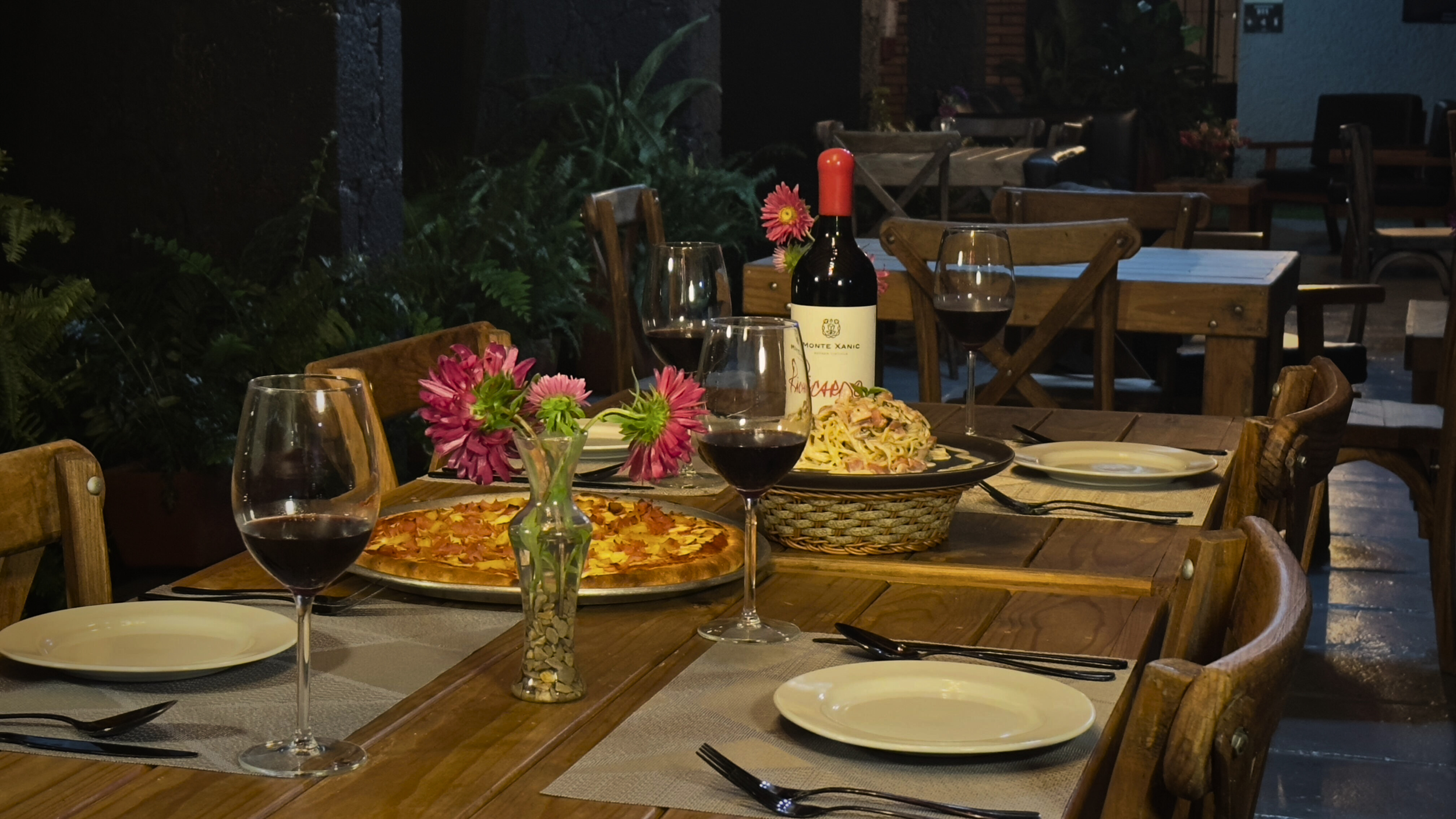 Mesa decorada con flores y comida como pizza y pasta, acompañada de copas de vino y una botella, en un ambiente al aire libre de noche.