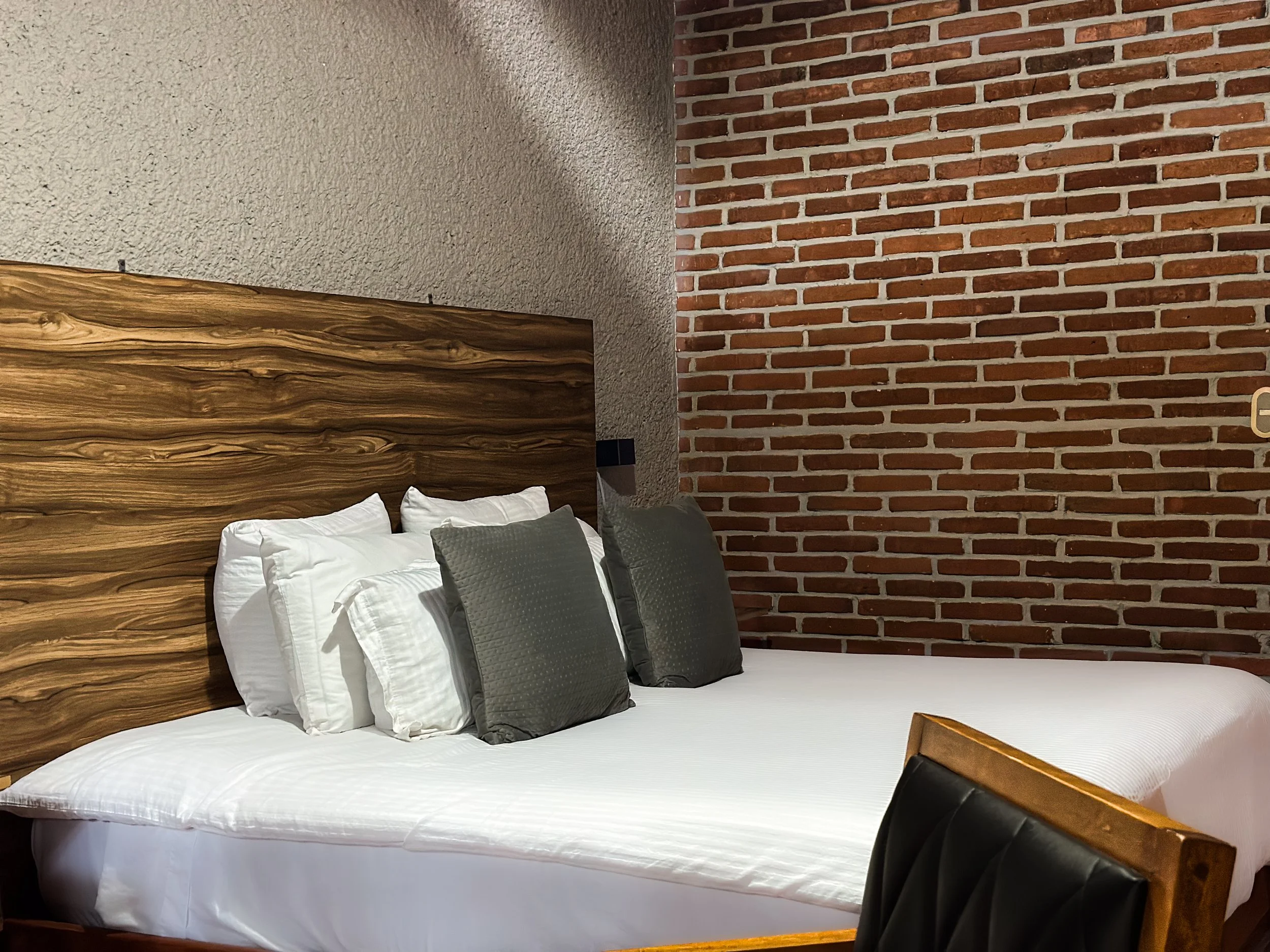 Cama con cabecera de madera y cuatro almohadas blancas y dos gris oscuras, en una habitación con paredes de cemento y ladrillo.