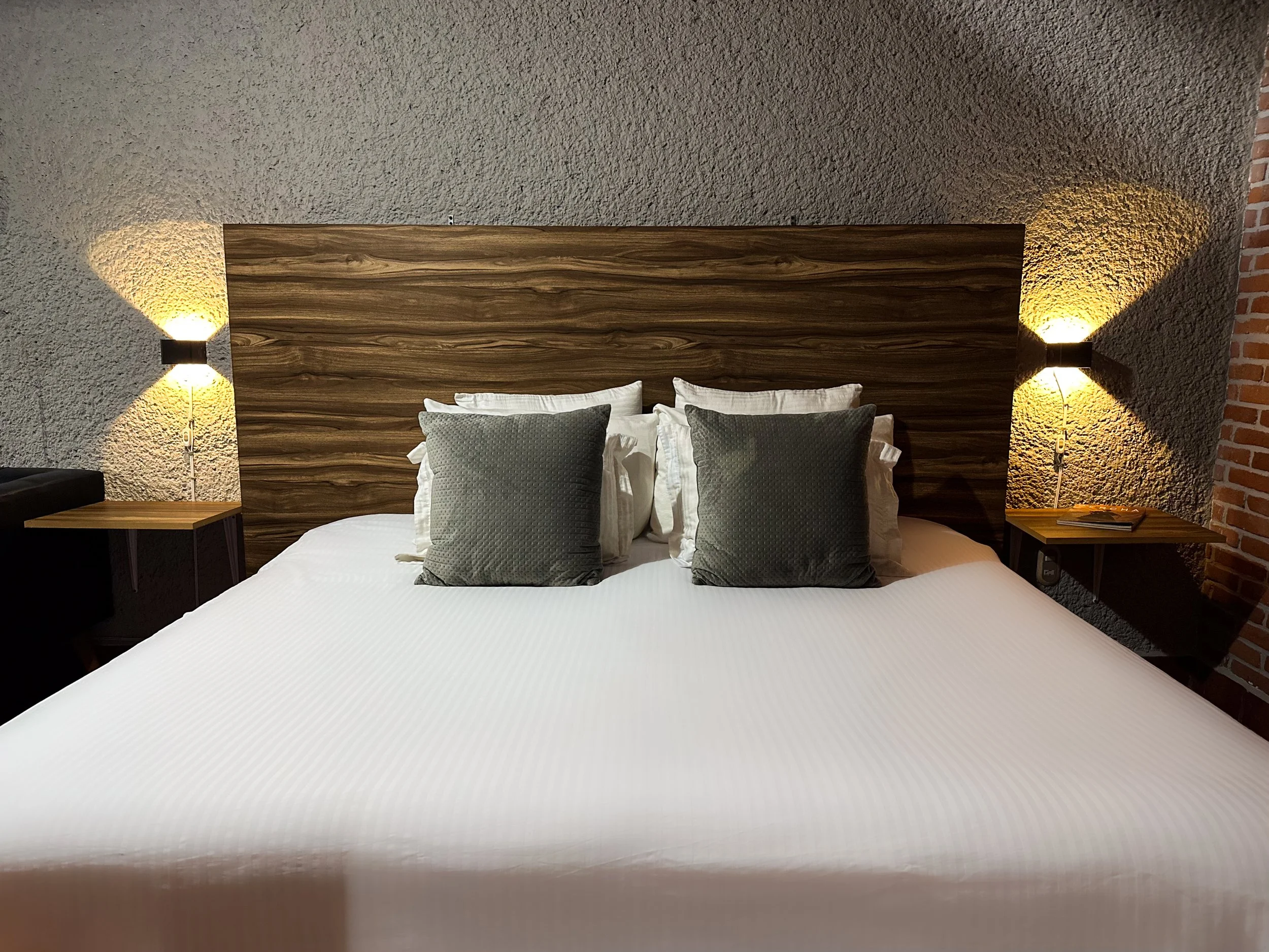 Cama con cabecera de madera, cuatro almohadas blancas y dos decorativas grises, lámparas de pared a cada lado, paredes de textura gris y ladrillo a la derecha.