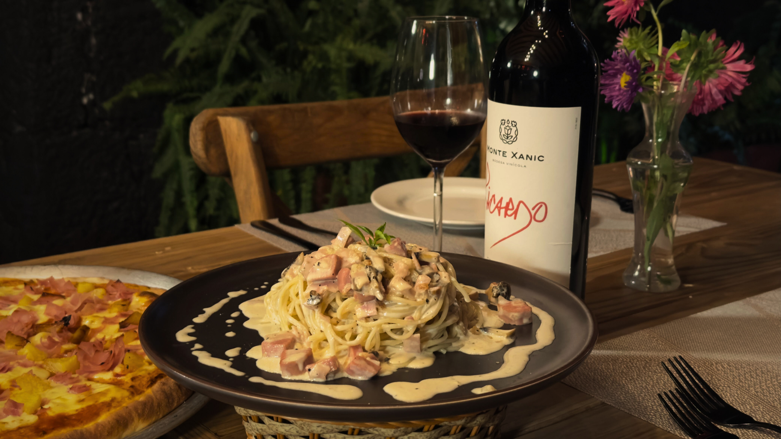 Cena con plato de pasta con salsa, pizza en la esquina, copa de vino tinto, botella de vino y flores en un florero en una mesa de madera.
