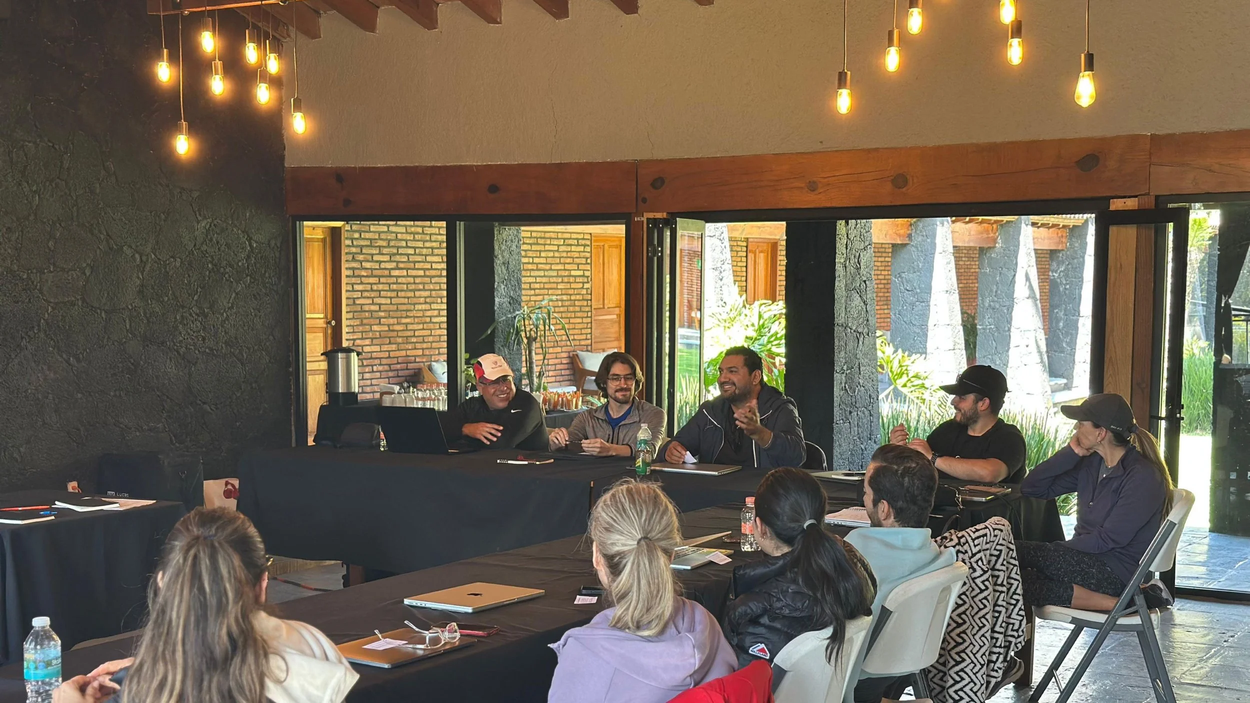Reunión de varias personas en una sala de conferencias, algunos participando en una discusión y otros escuchando, con muebles y ventanas grandes que muestran un entorno exterior natural con árboles y plantas.